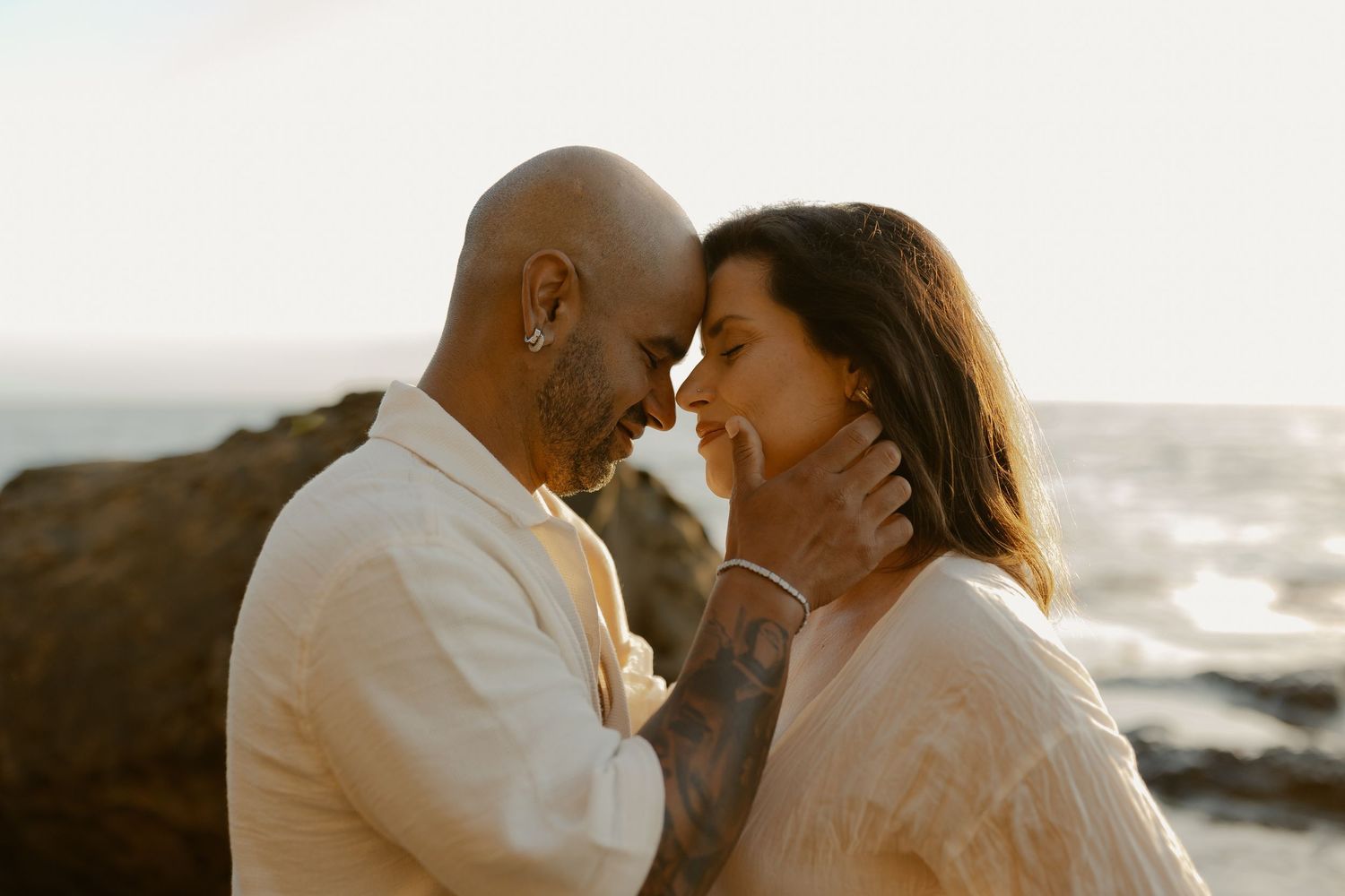 A romantic couple shares an intimate moment at sunset on a rocky coastal overlook with ocean waves in the background.