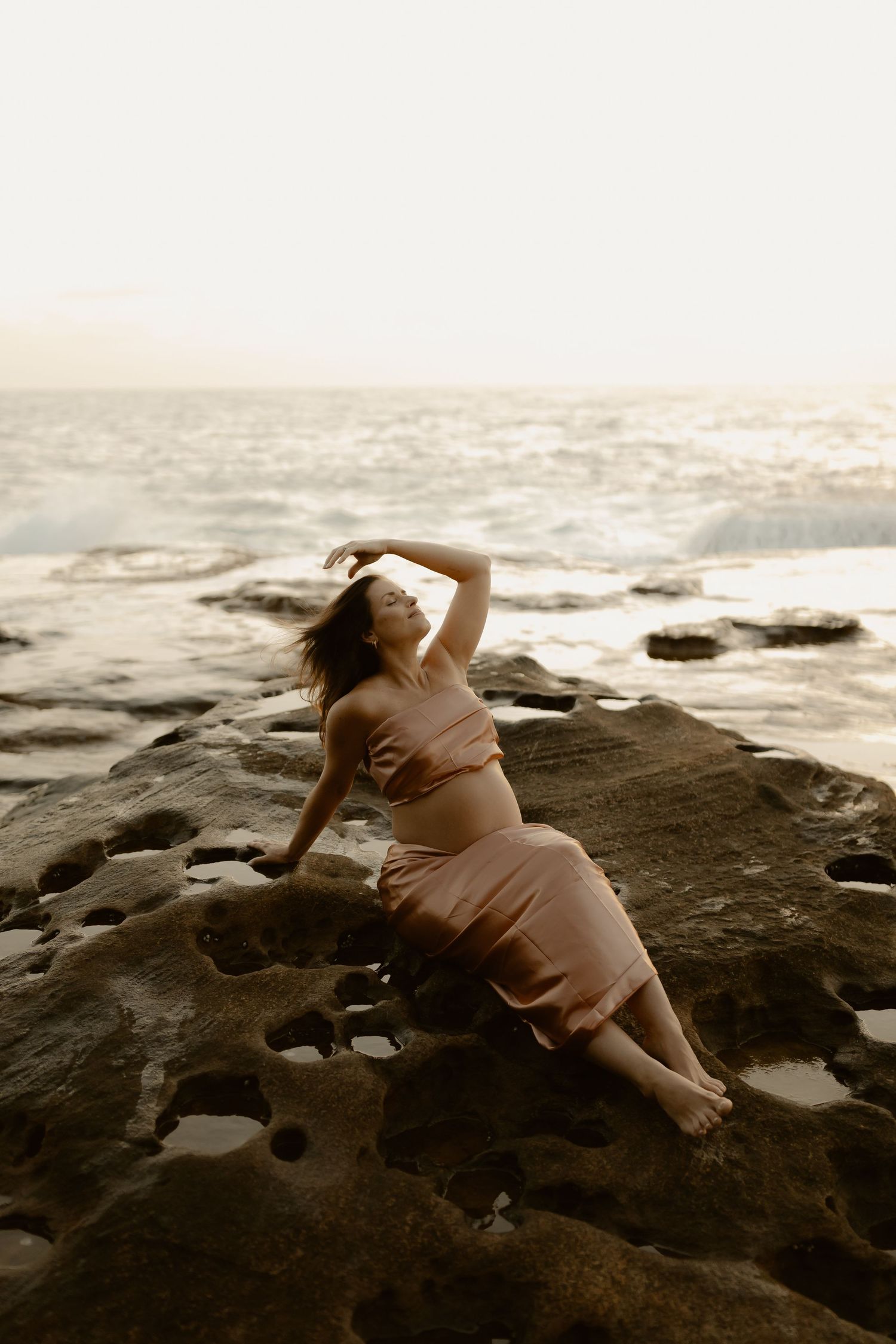 A person in a pink dress reclines on coastal rocks at sunset with dramatic ocean waves in the background.