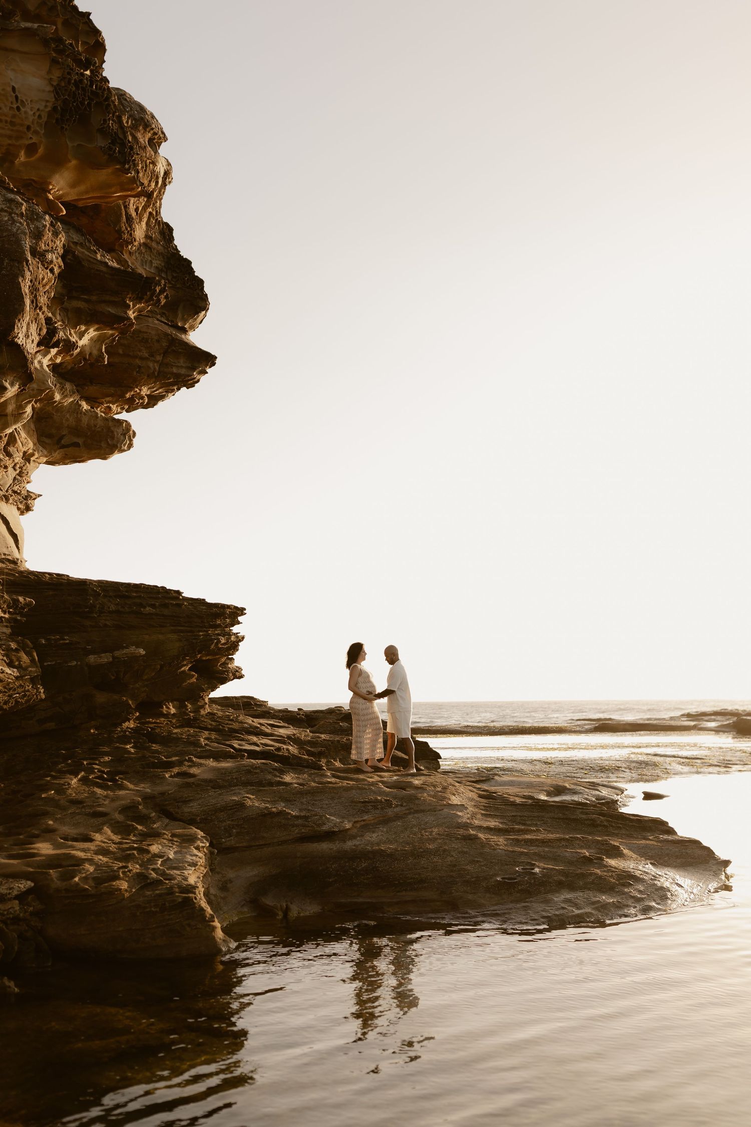 Two figures stand together on a dramatic rocky cliff overlooking the ocean at sunset in a sepia-toned photograph.