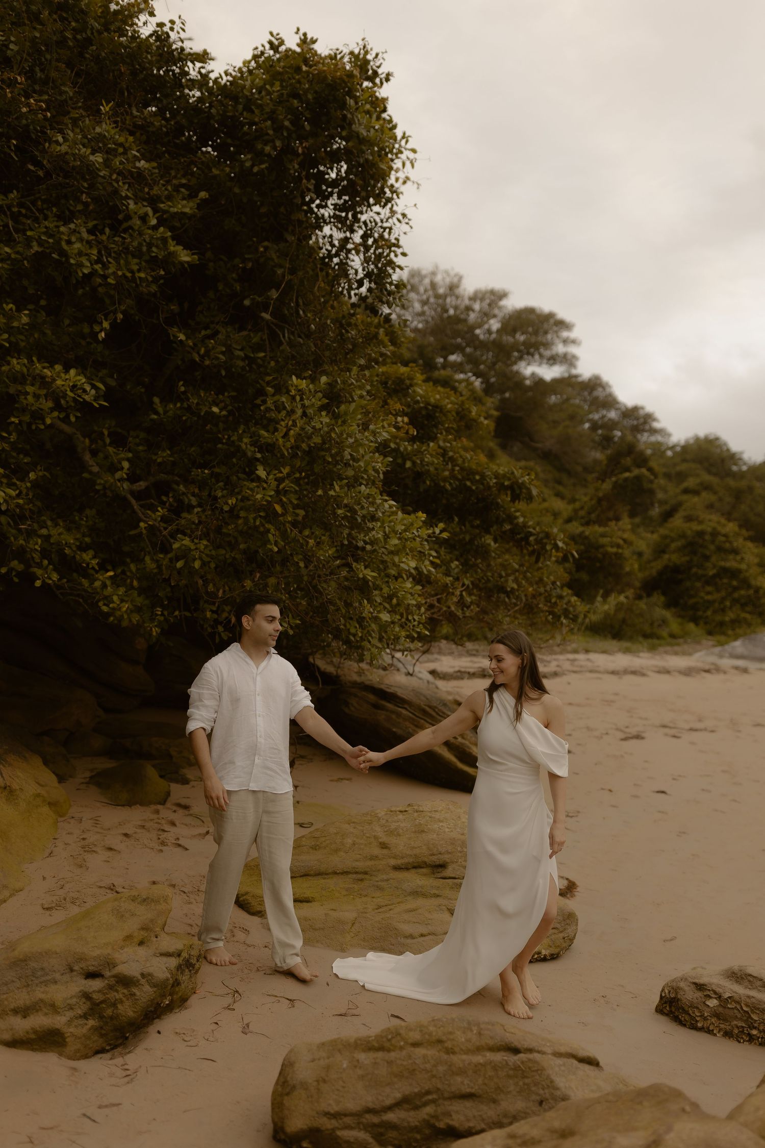 A couple dances together on a sandy beach with lush greenery in the background.