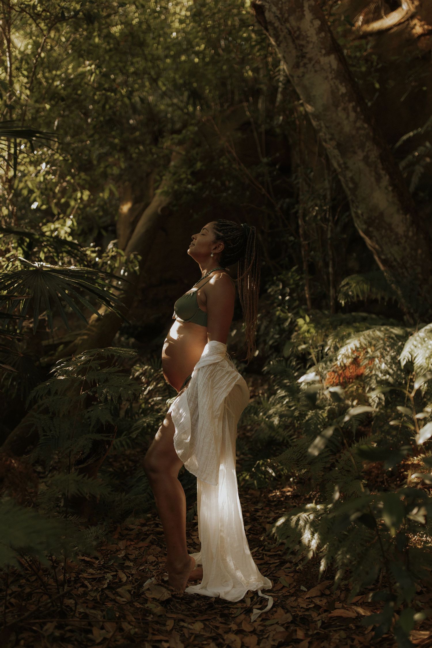 Artistic silhouette of an expectant mother in flowing white dress standing in dappled forest light.