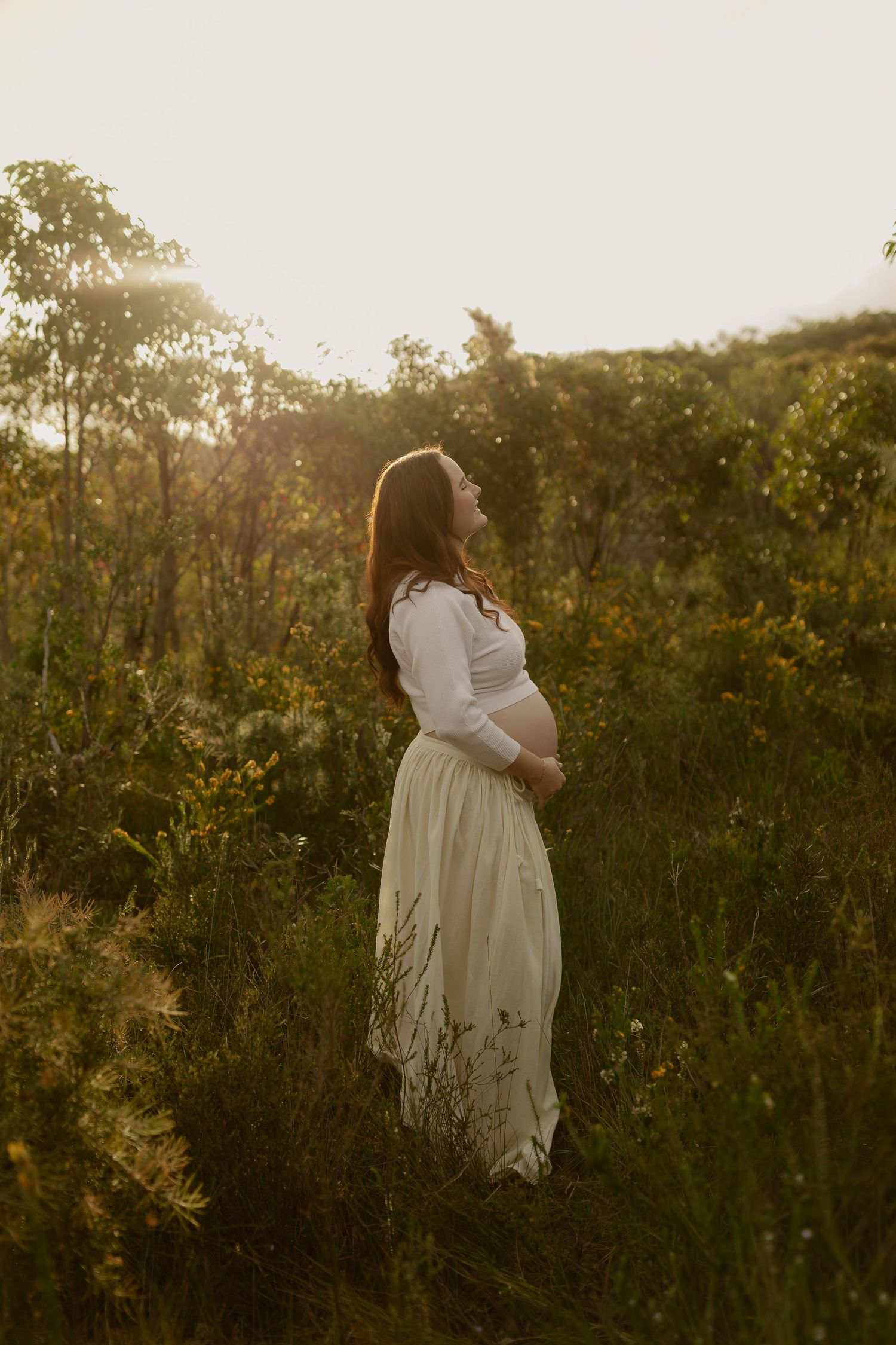 Pregnant woman in white dress standing in golden sunlit meadow, cradling baby bump surrounded by wildflowers at sunset.