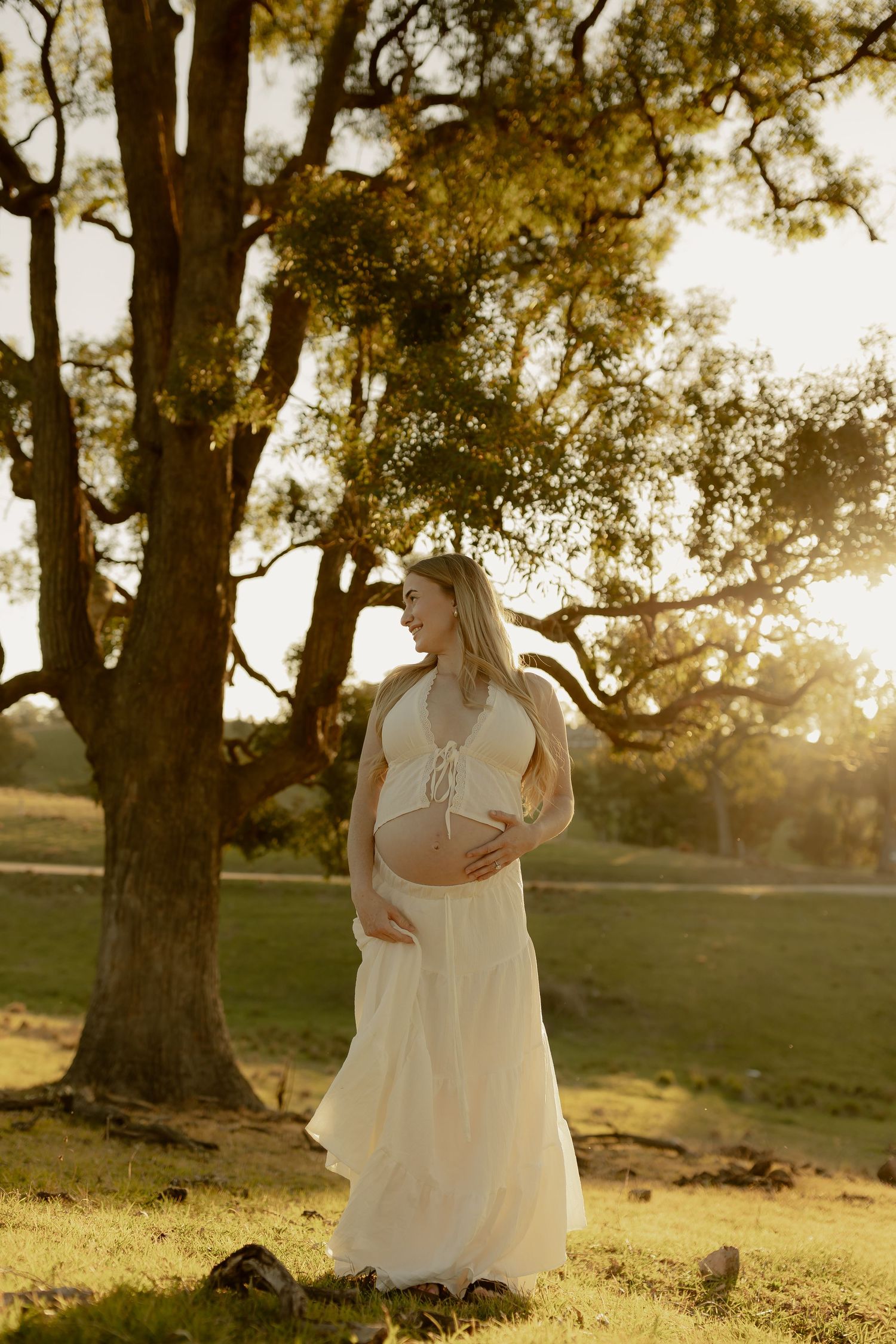 Pregnant woman in white crop top and skirt cradling baby bump outdoors at golden hour near large oak tree.