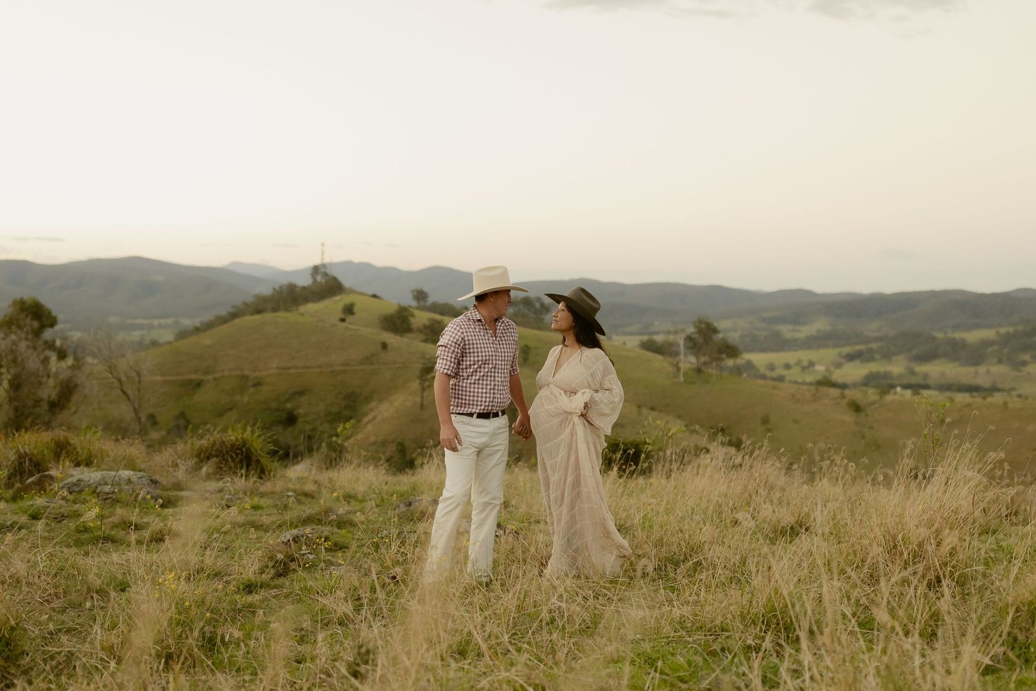Couple standing on a scenic hilltop in golden hour light, man in cowboy hat, woman in flowing dress.