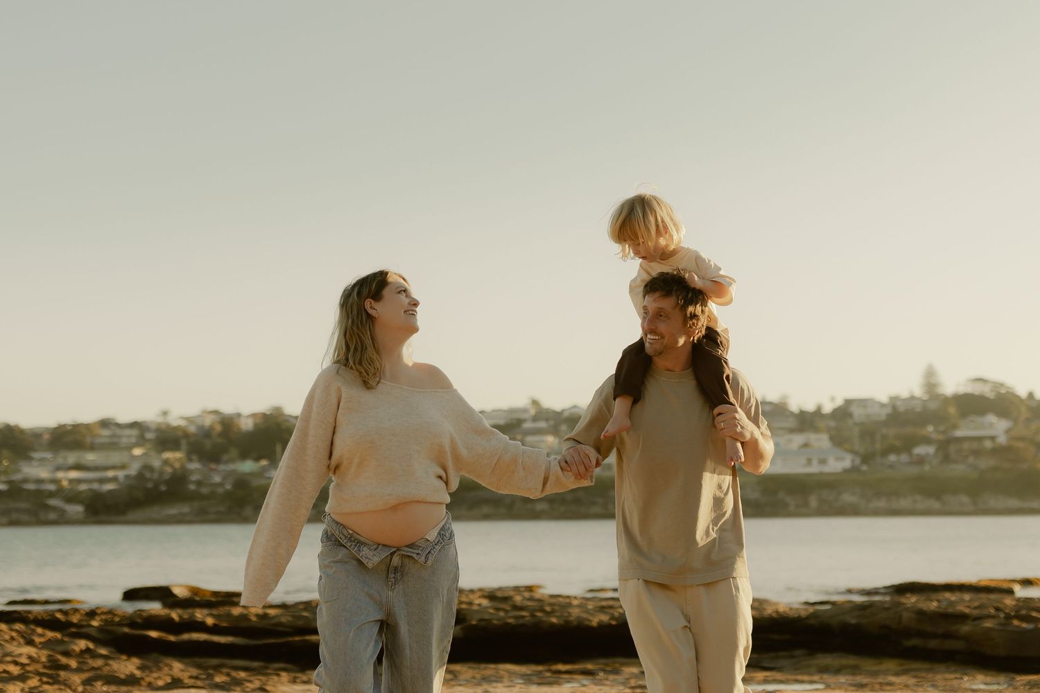 Two women and a child enjoy a sunny beach day, one carrying the toddler on her shoulders near the water.