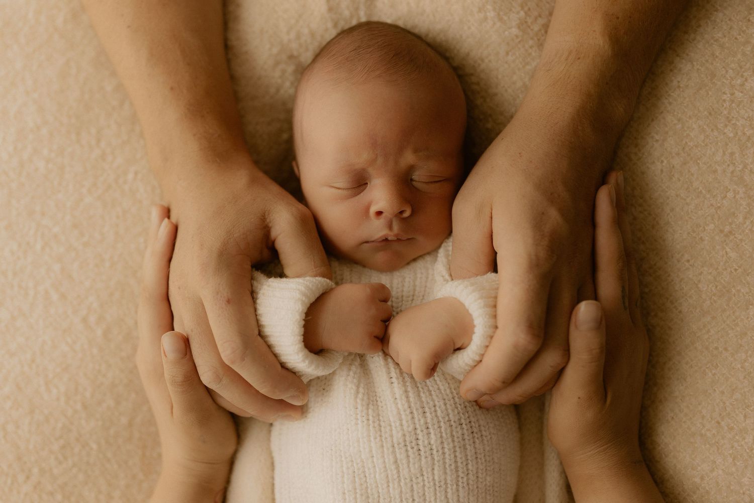 Gentle hands cradling a sleeping newborn wrapped in white knit fabric.