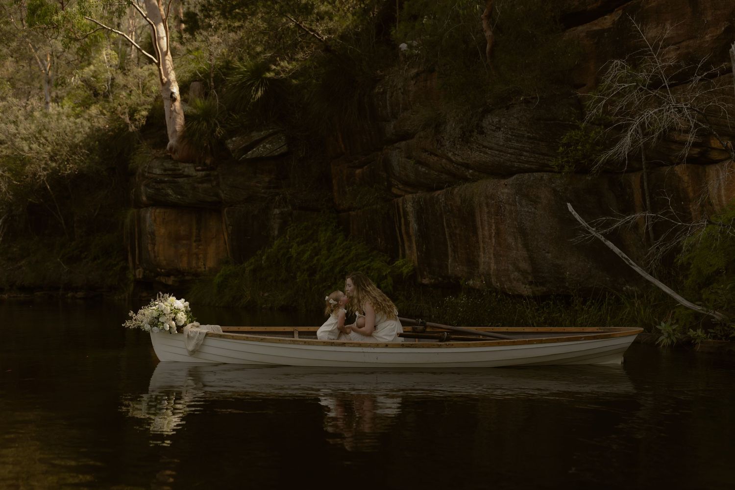 A romantic scene with two people sitting in a white rowboat on a dark, moody lake surrounded by rocky cliffs.