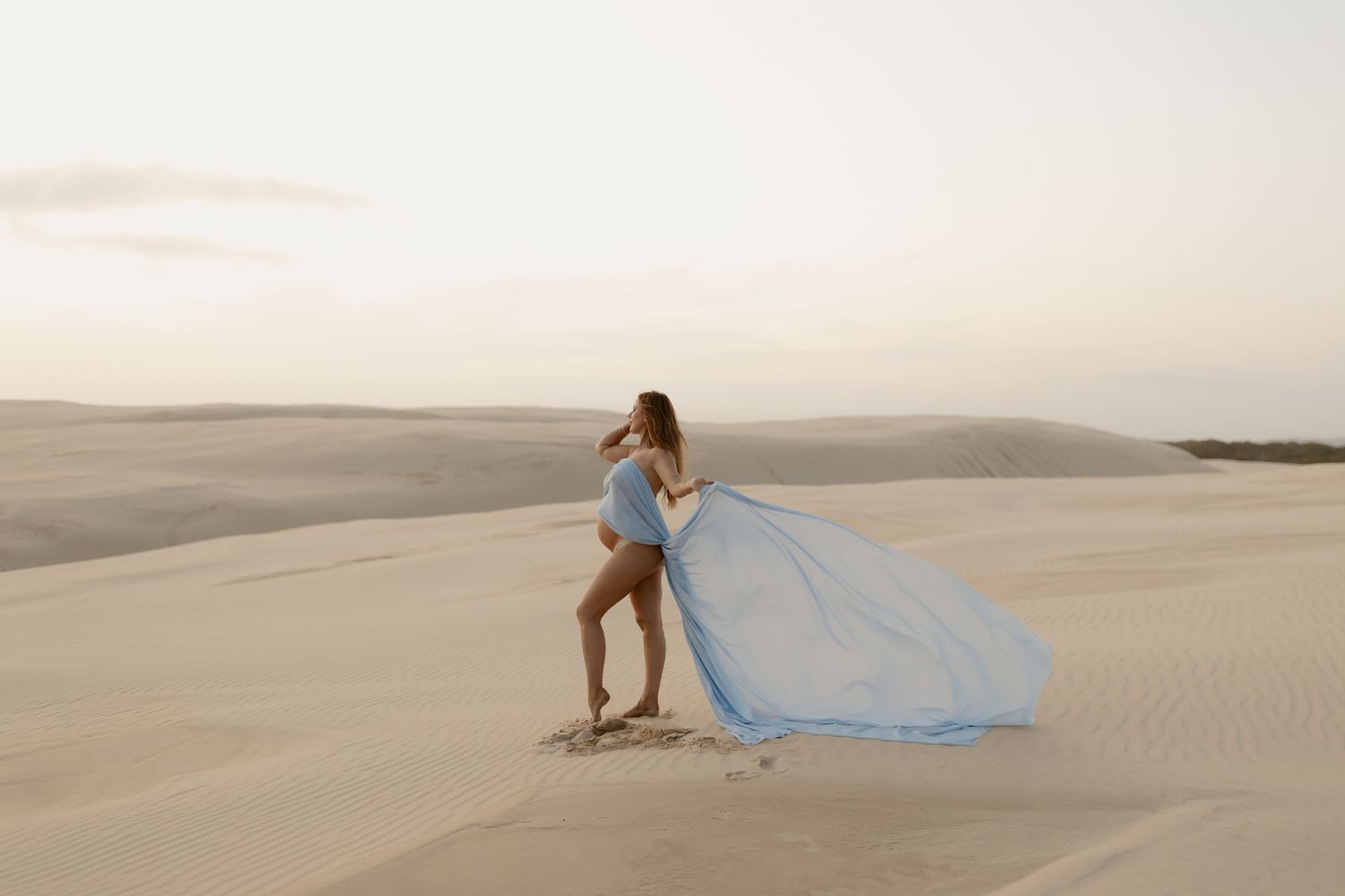 A flowing light blue dress billows in the wind as someone poses on desert sand dunes at sunset.