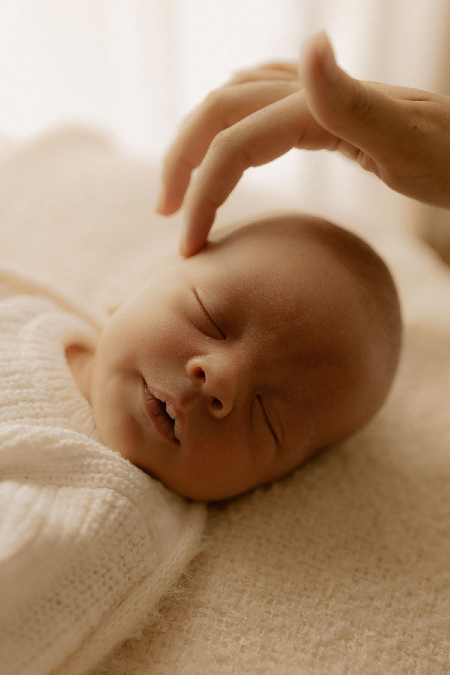 A gentle hand touches the head of a peaceful sleeping newborn baby swaddled in white knit fabric.