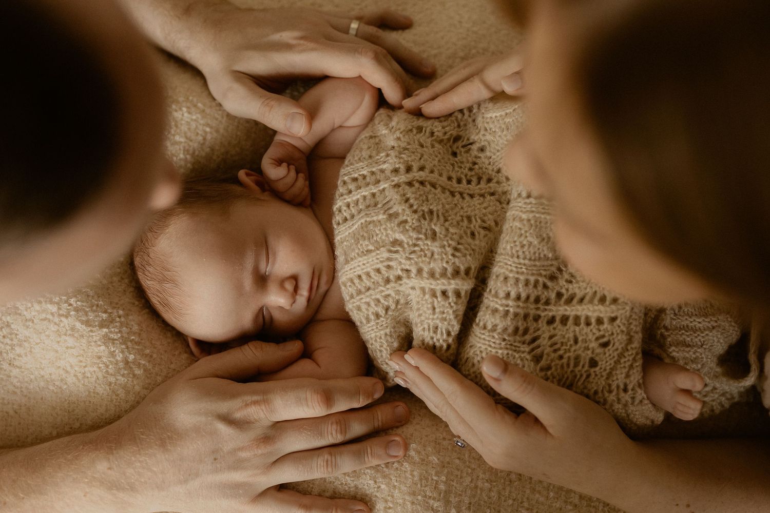 A baby wearing a cream knit outfit lies peacefully while being held by gentle hands in a warm sepia-toned photograph.