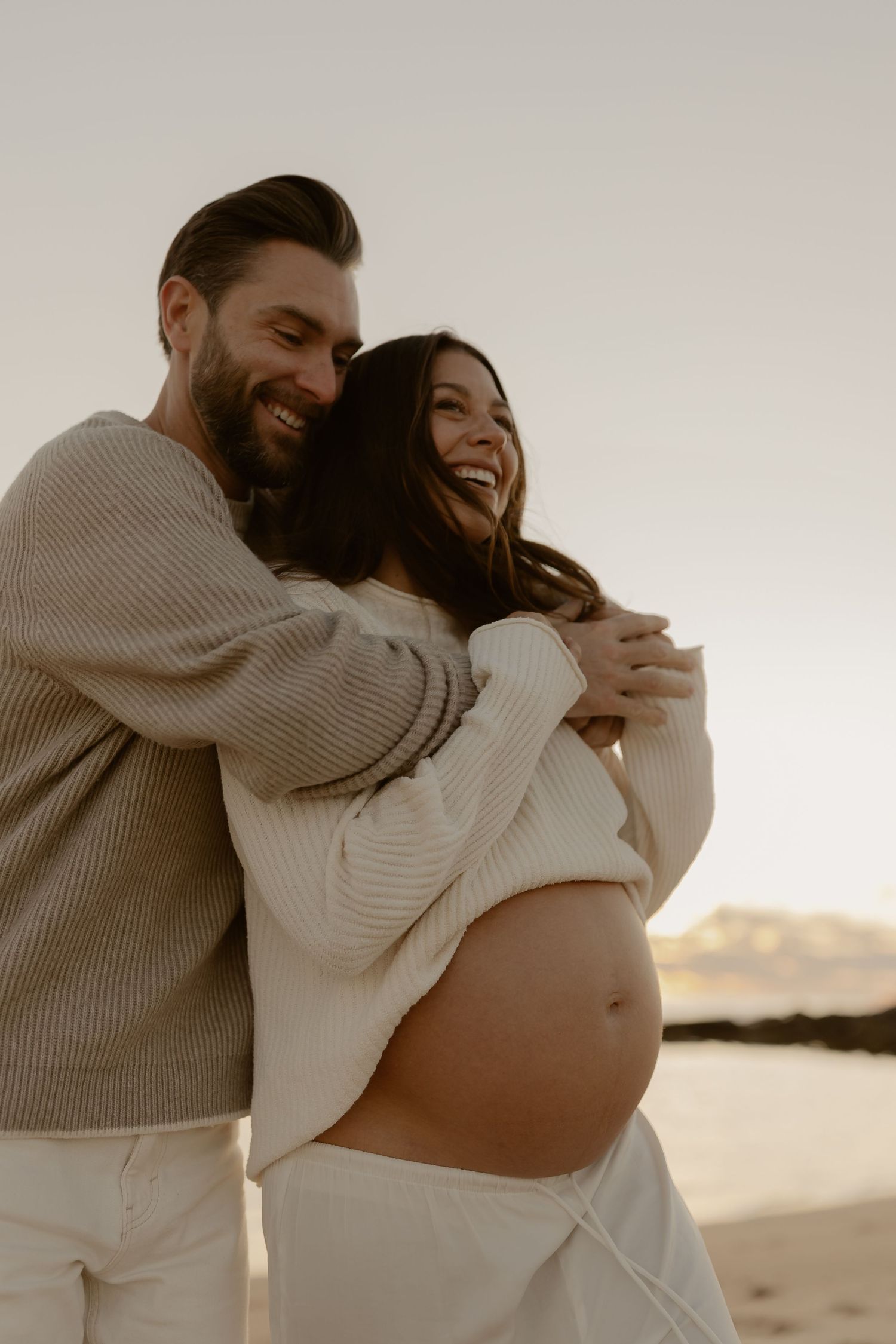 A couple embraces on a beach at sunset while celebrating a pregnancy in matching beige sweaters.