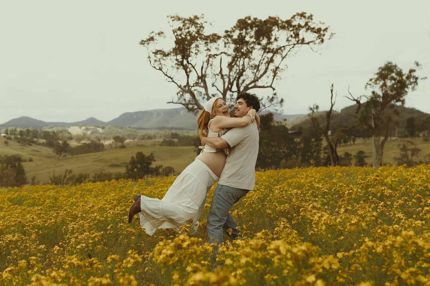 A couple shares an intimate moment while standing among bright yellow wildflowers in a rustic countryside setting.