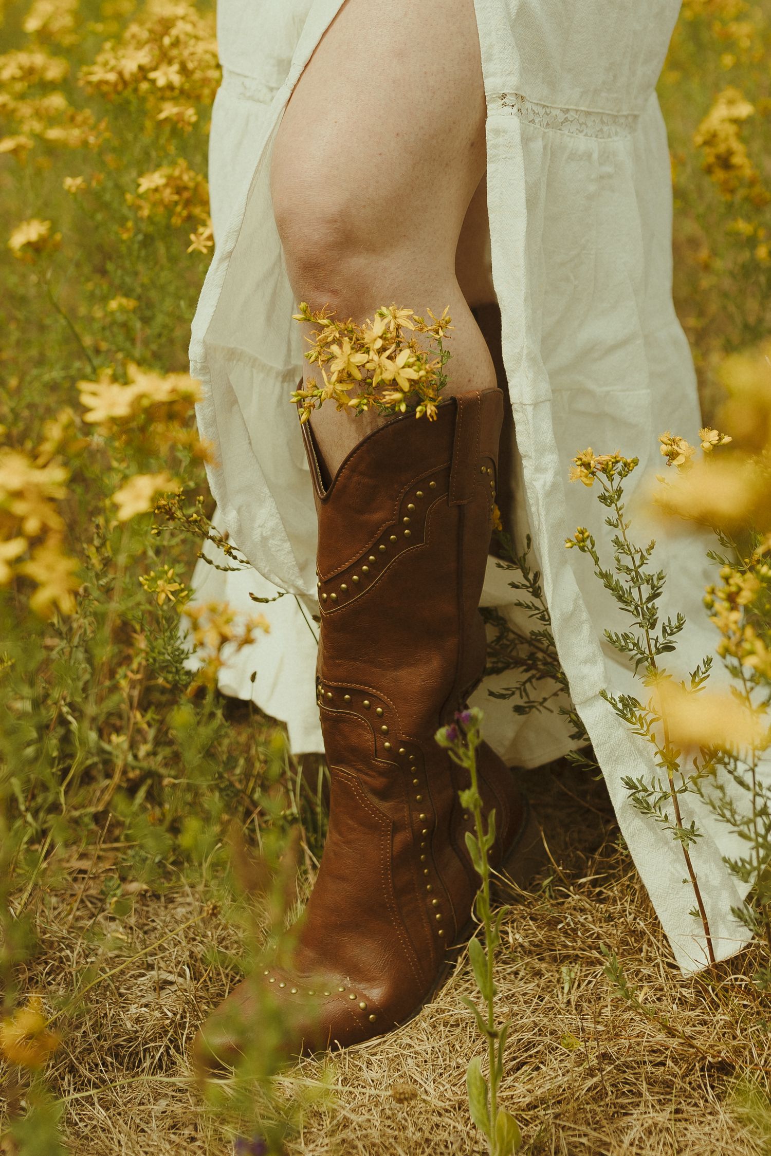 Brown leather cowboy boots stand in contrast against a white dress hem amid yellow wildflowers in golden light.