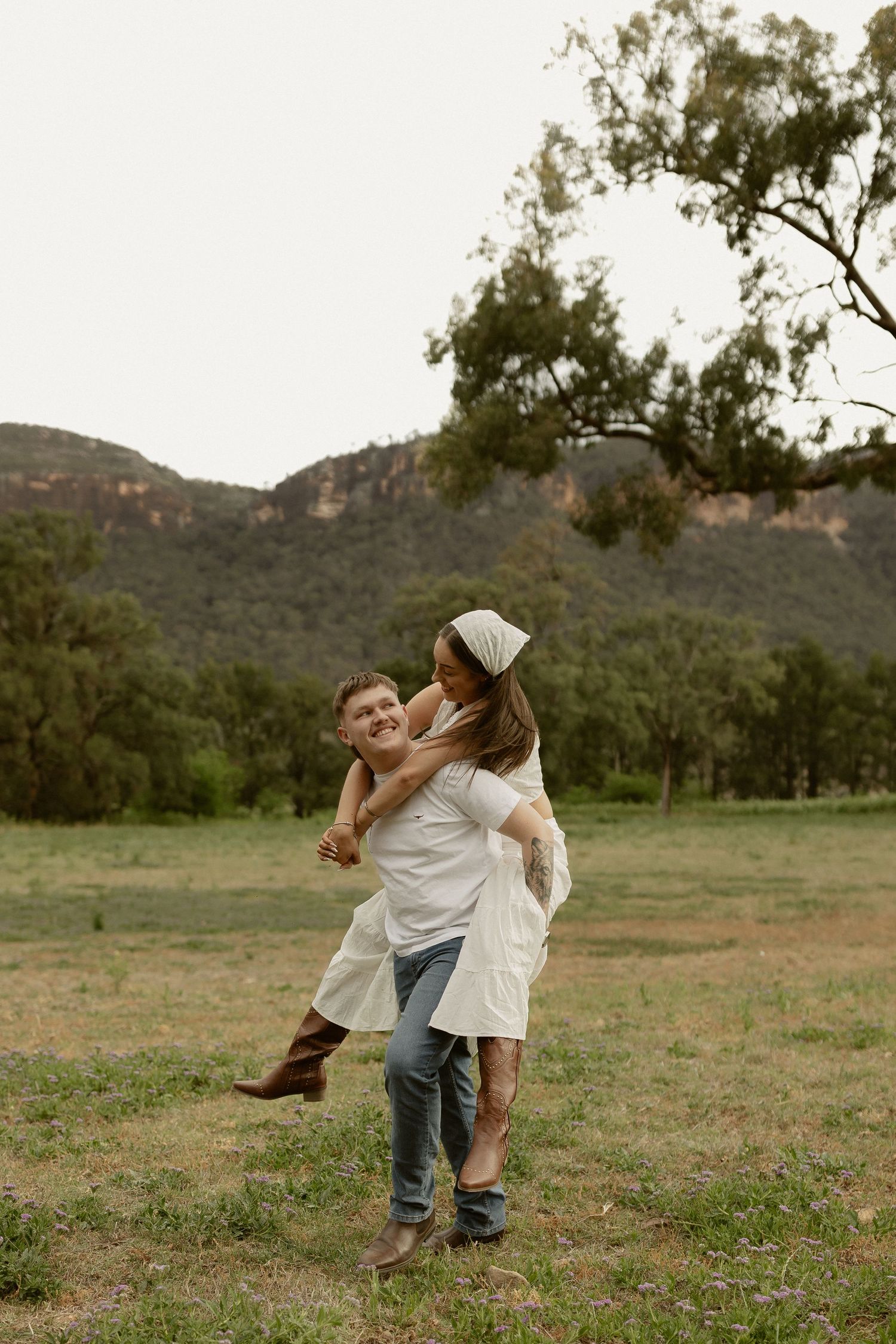 A couple dances playfully in a grassy field with mountains and trees in the background.