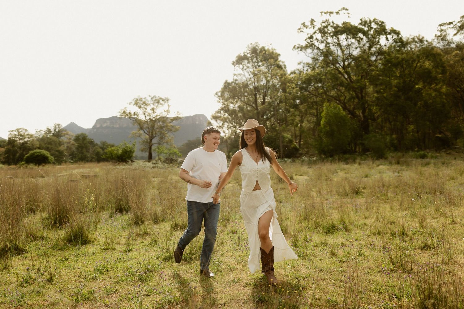 Two people run playfully through a golden meadow with mountains and trees in the background during sunset.