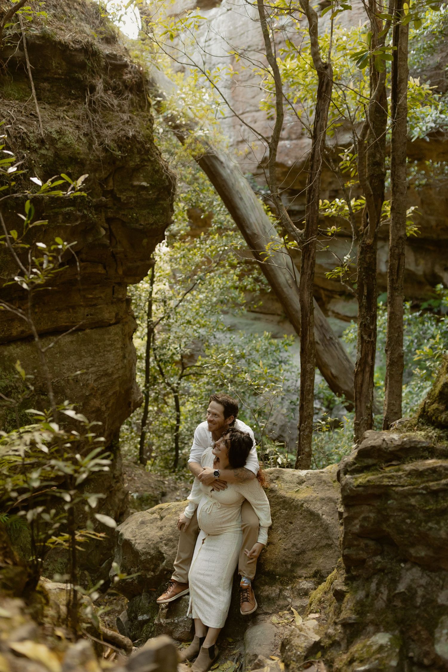 Couple exploring rocky cliffs and caves in a lush forest setting during golden hour.