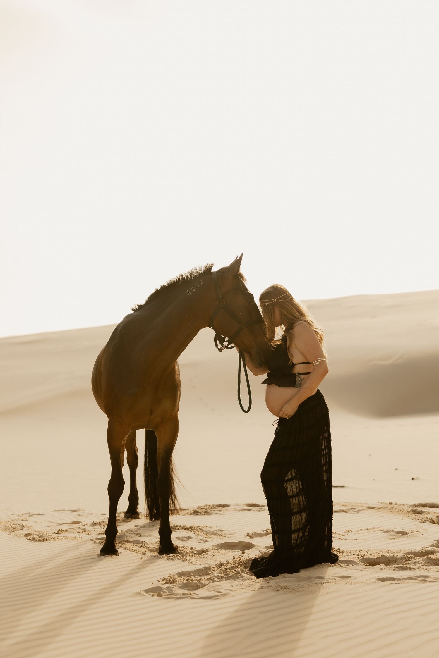A person in dark clothing kneels beside a black horse on sandy desert dunes at sunset.