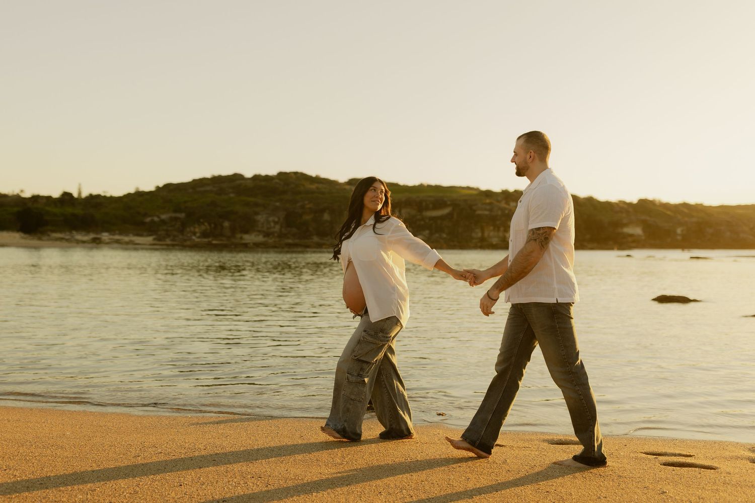 Pregnant couple holding hands walking on a golden sandy beach at sunset with calm water in the background.