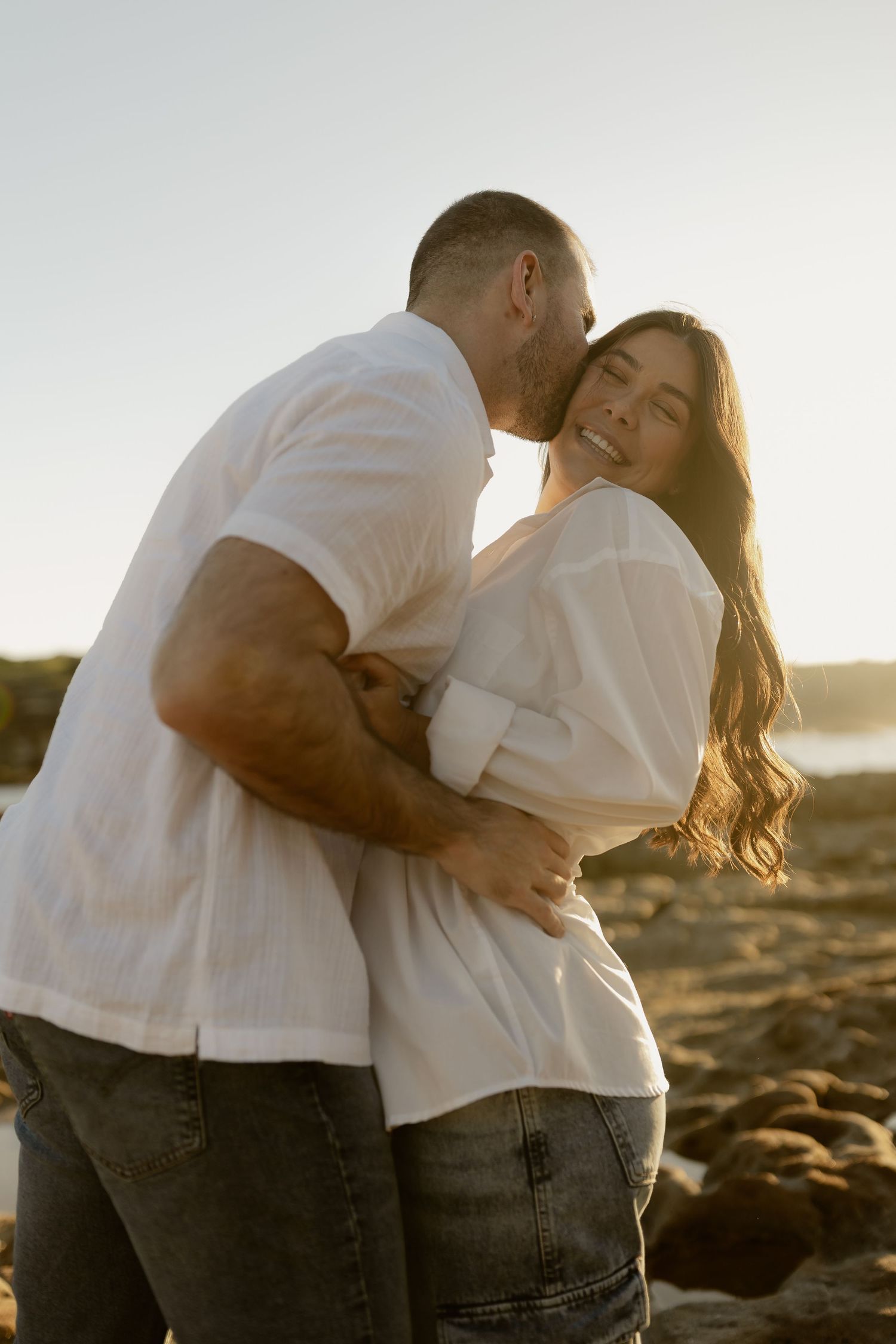 Couple embracing at golden hour on a rocky beach, man kissing pregnant woman's cheek as she smiles.