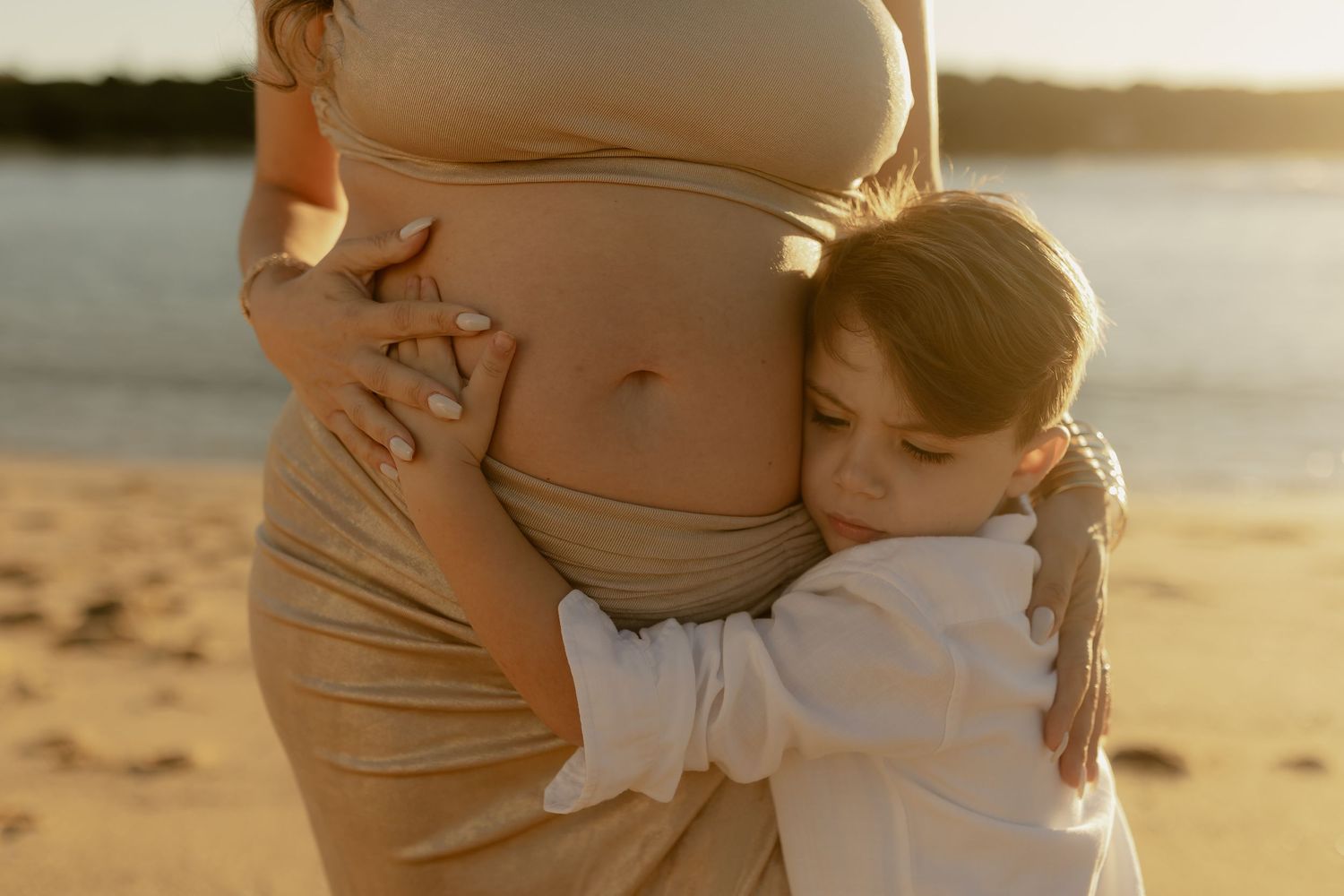 A young child hugging a pregnant woman's belly on a sunny beach at golden hour.