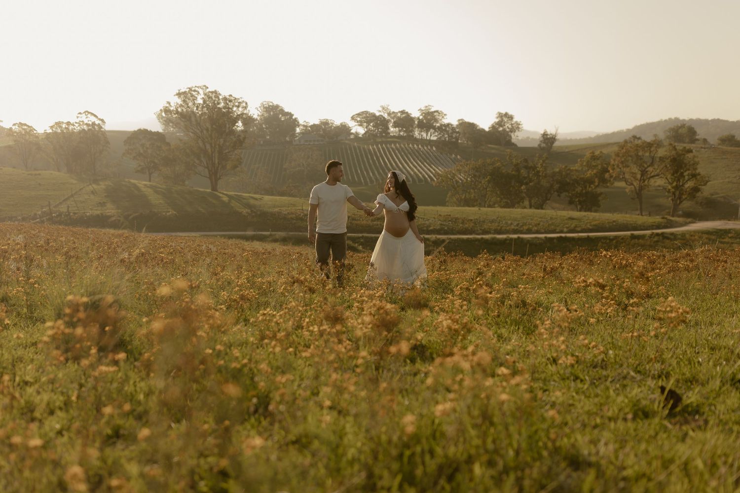 A couple walks hand in hand through a golden wildflower meadow at sunset with rolling hills in the background.