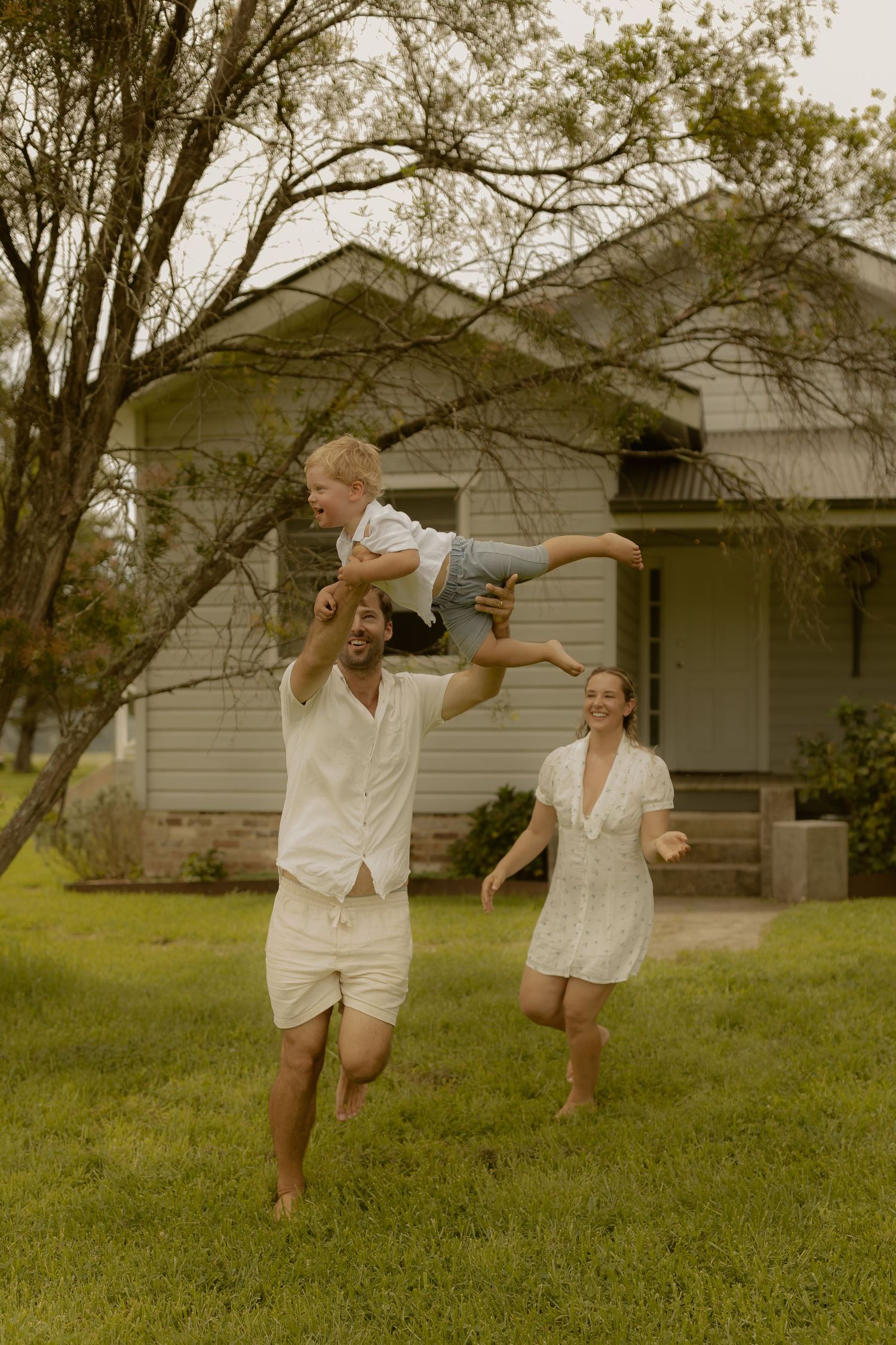 People in white outfits dance and play in front of a rustic house surrounded by trees.