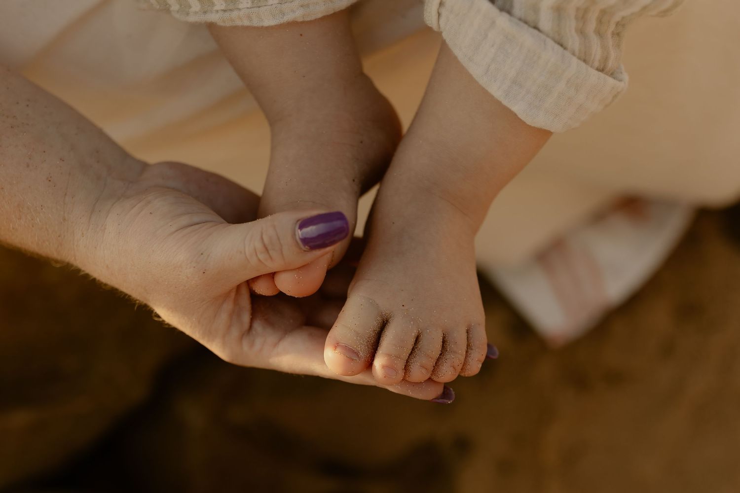 A gentle hand with purple nail polish tenderly holds a small baby's foot against a warm brown background.