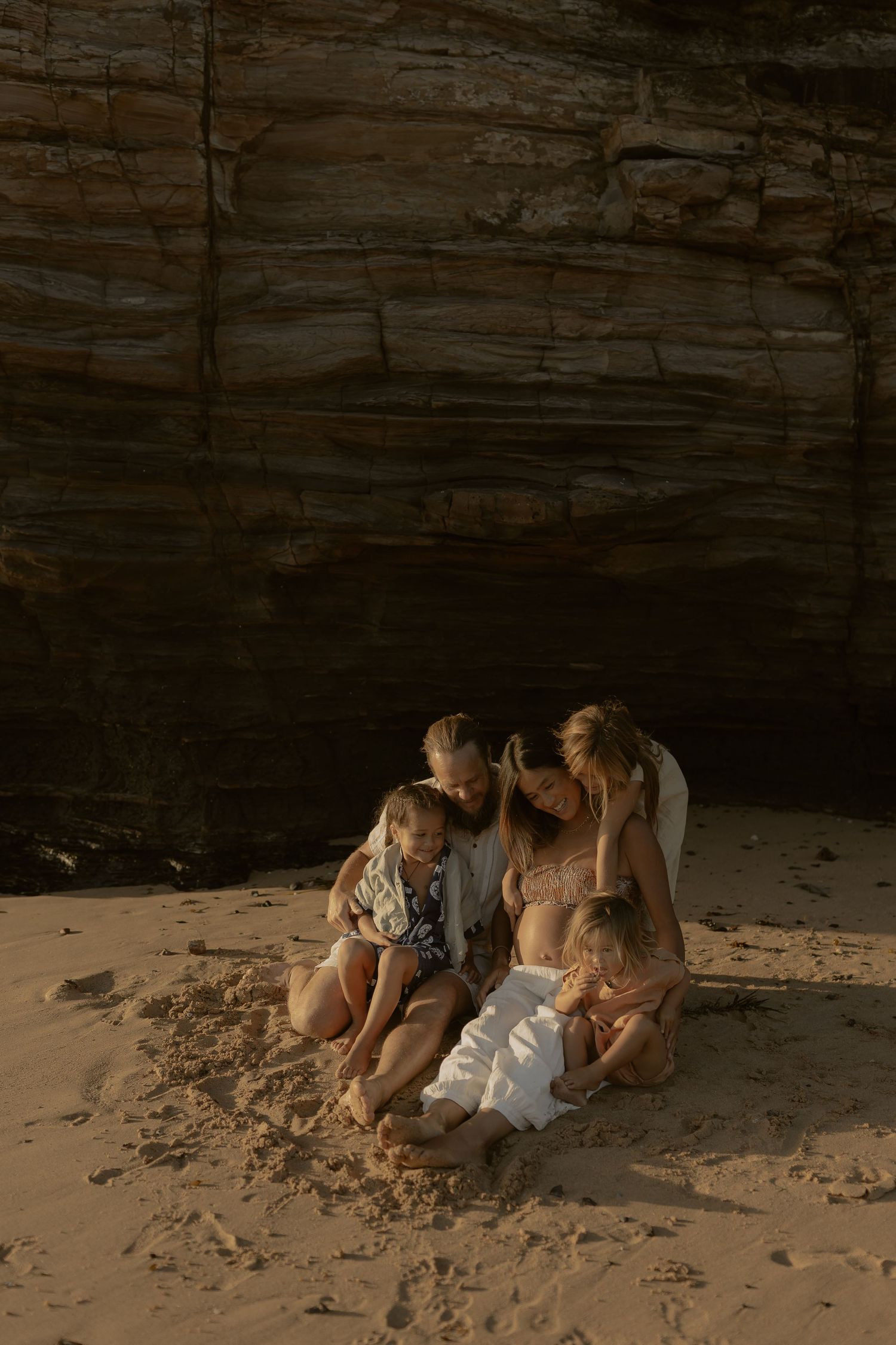A family sits together on a sandy beach against dramatic rocky cliffs during golden hour.
