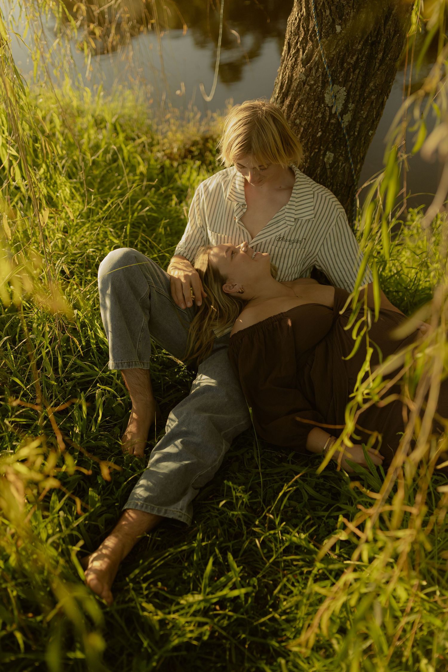 Couple resting in golden sunlight by a lake, leaning against a tree trunk surrounded by lush green grass.