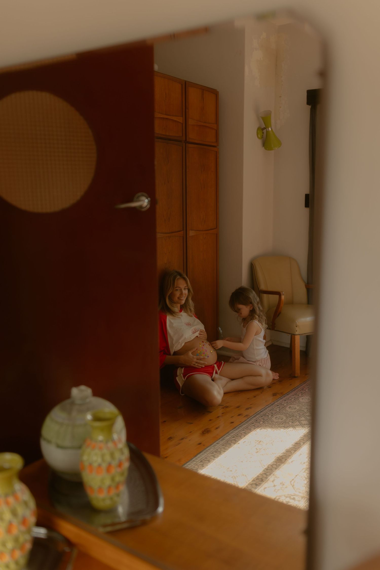 A pregnant woman sits on the floor with a young child in a warm, sunlit room reflected in a mirror.