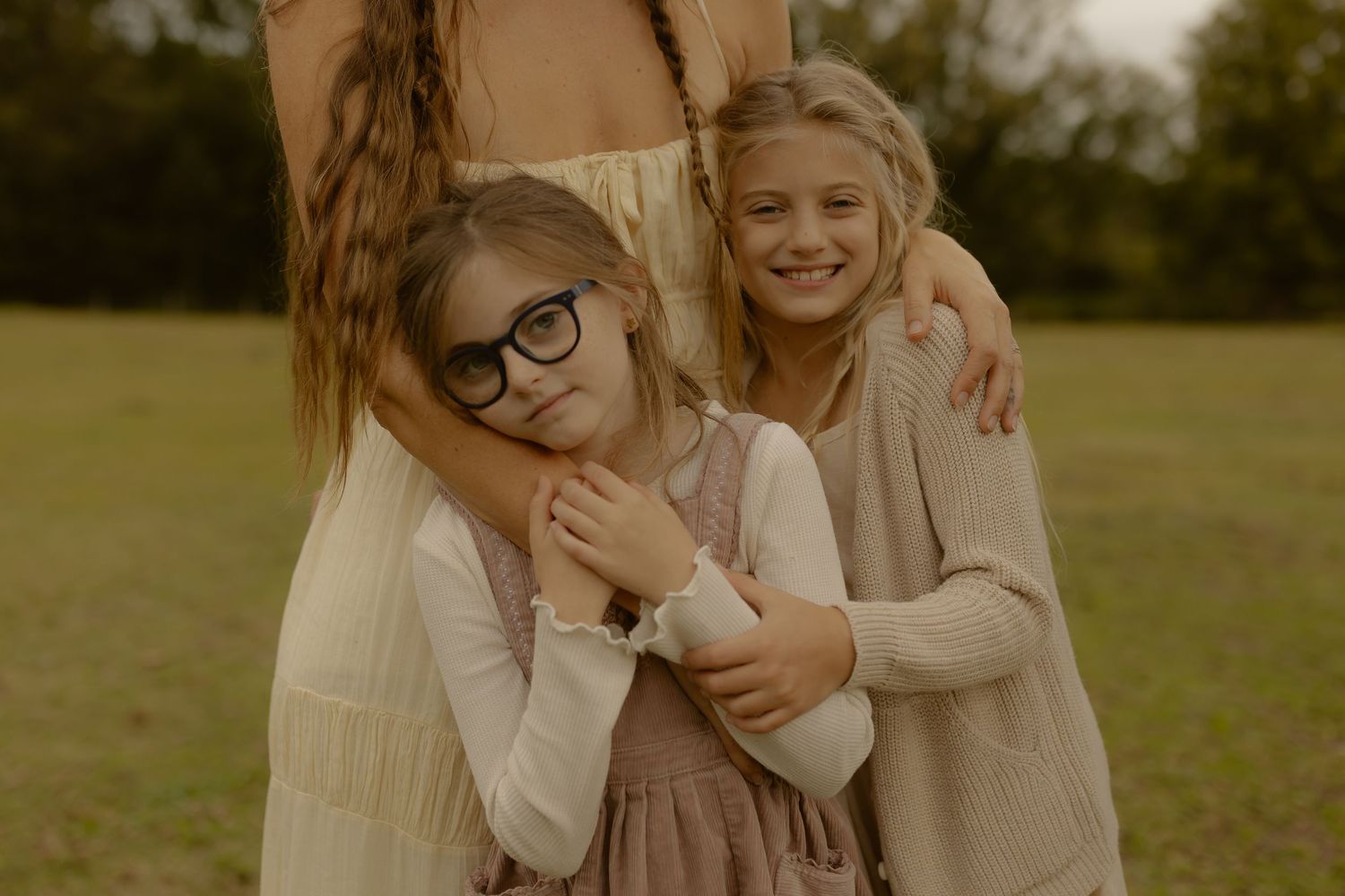 Two blonde sisters hug their mother outdoors in a green park setting.