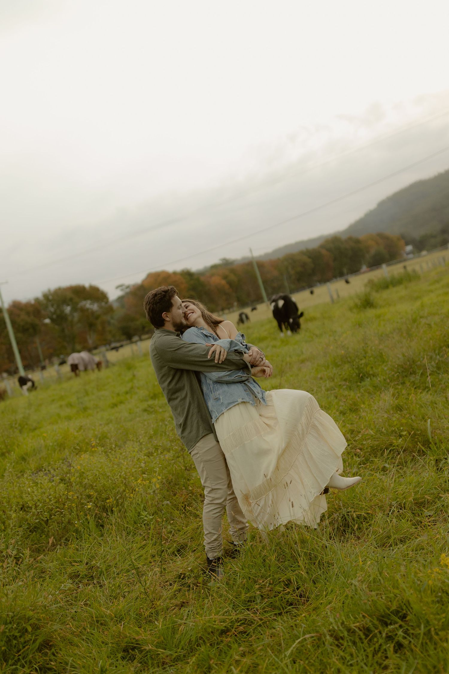 Couple embracing and dancing in a lush green field with cows grazing in the background on a cloudy day.