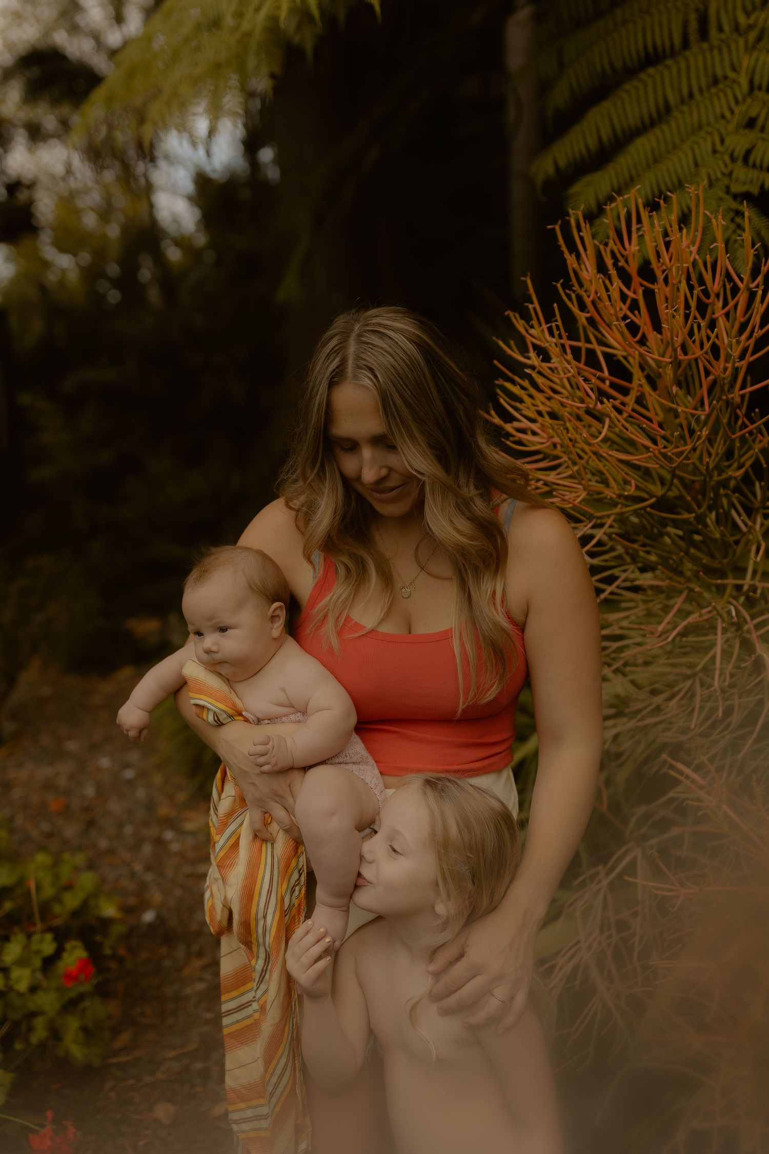 A mother in a red top holds her baby while her toddler leans in, surrounded by lush garden greenery.