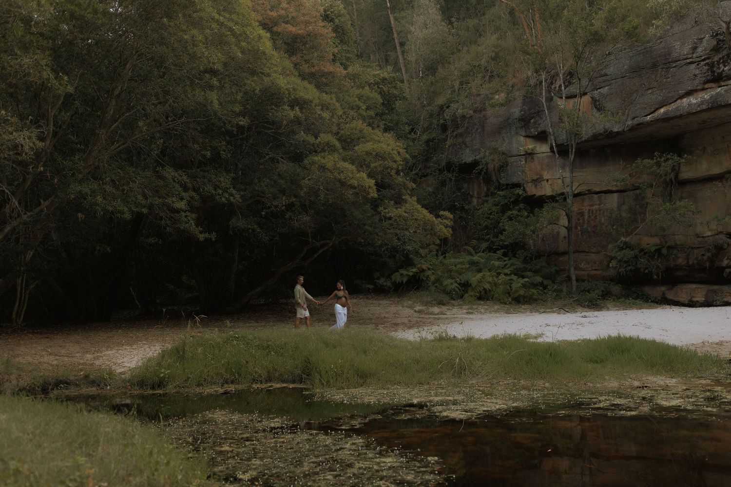 Two people fishing along a serene river bank surrounded by lush green trees and towering sandstone cliffs.