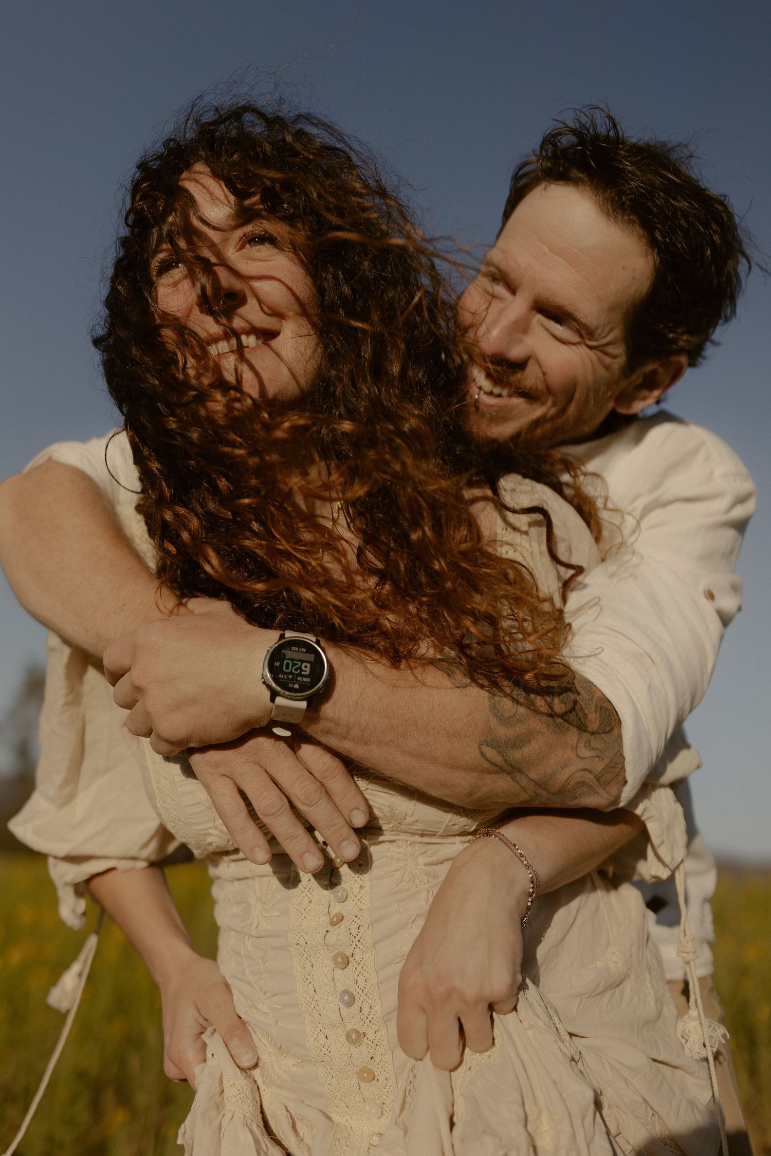 A joyful couple embraces outdoors, man in white shirt wearing a smartwatch hugs curly-haired woman in a sunny field.