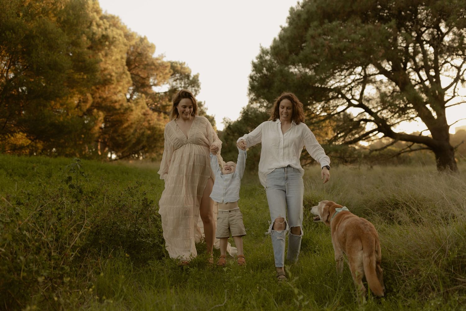 Family wearing coordinated beige and white outfits poses together in a golden sunset field during outdoor photo session.