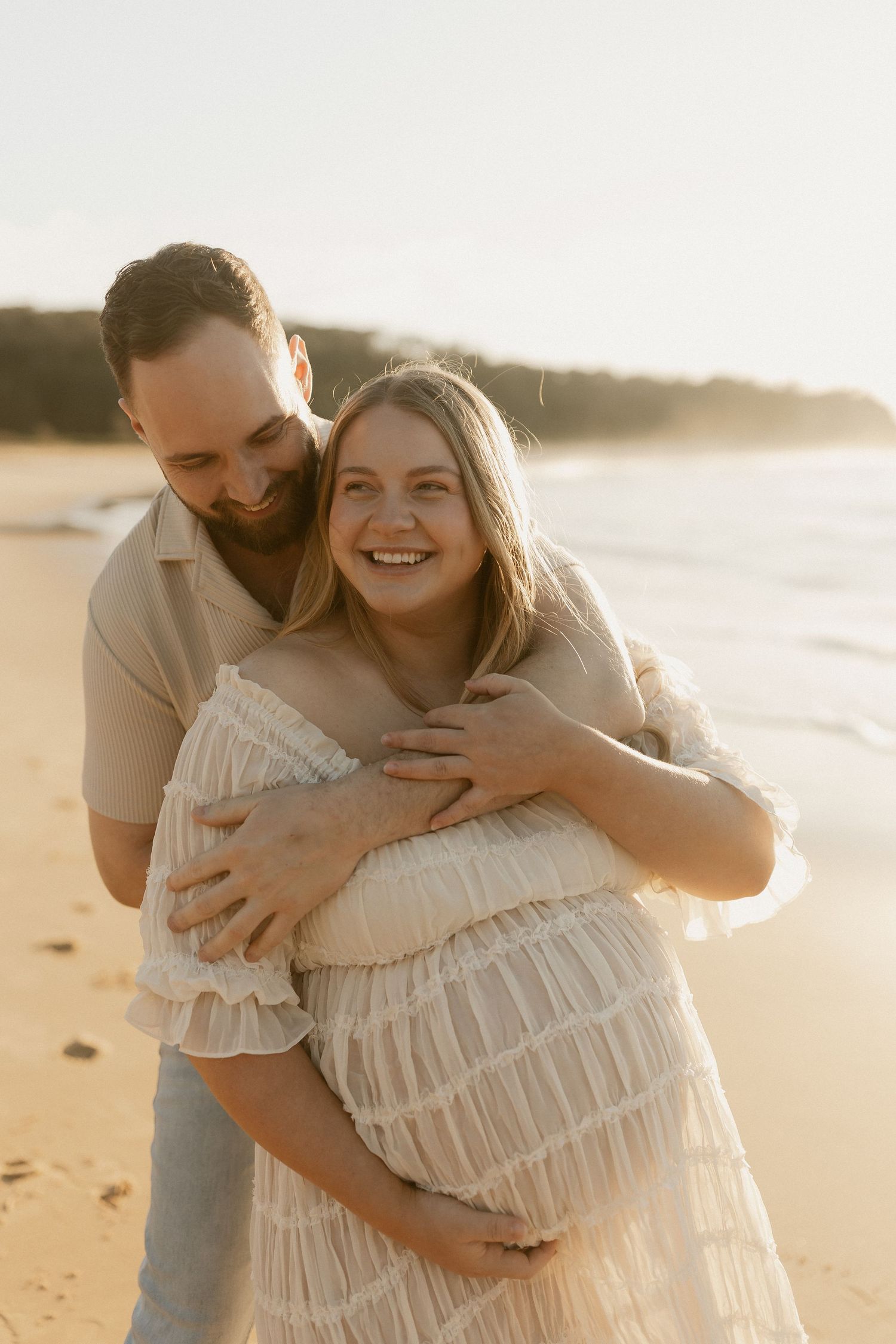 Couple embracing on a sandy beach at golden hour, pregnant woman in white lace dress smiling joyfully.