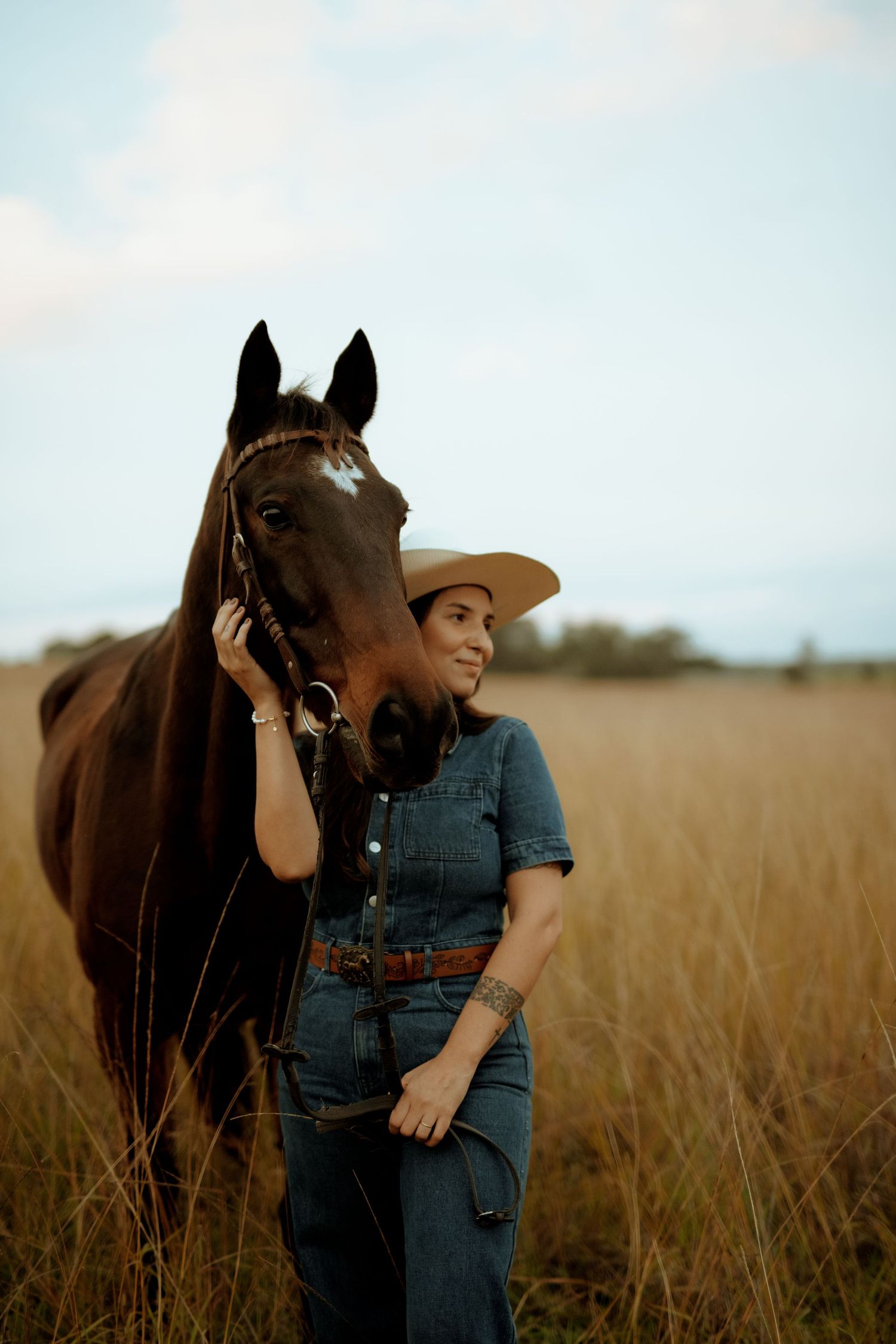 A smiling woman in denim overalls and cowboy hat stands beside a dark brown horse in a golden field.