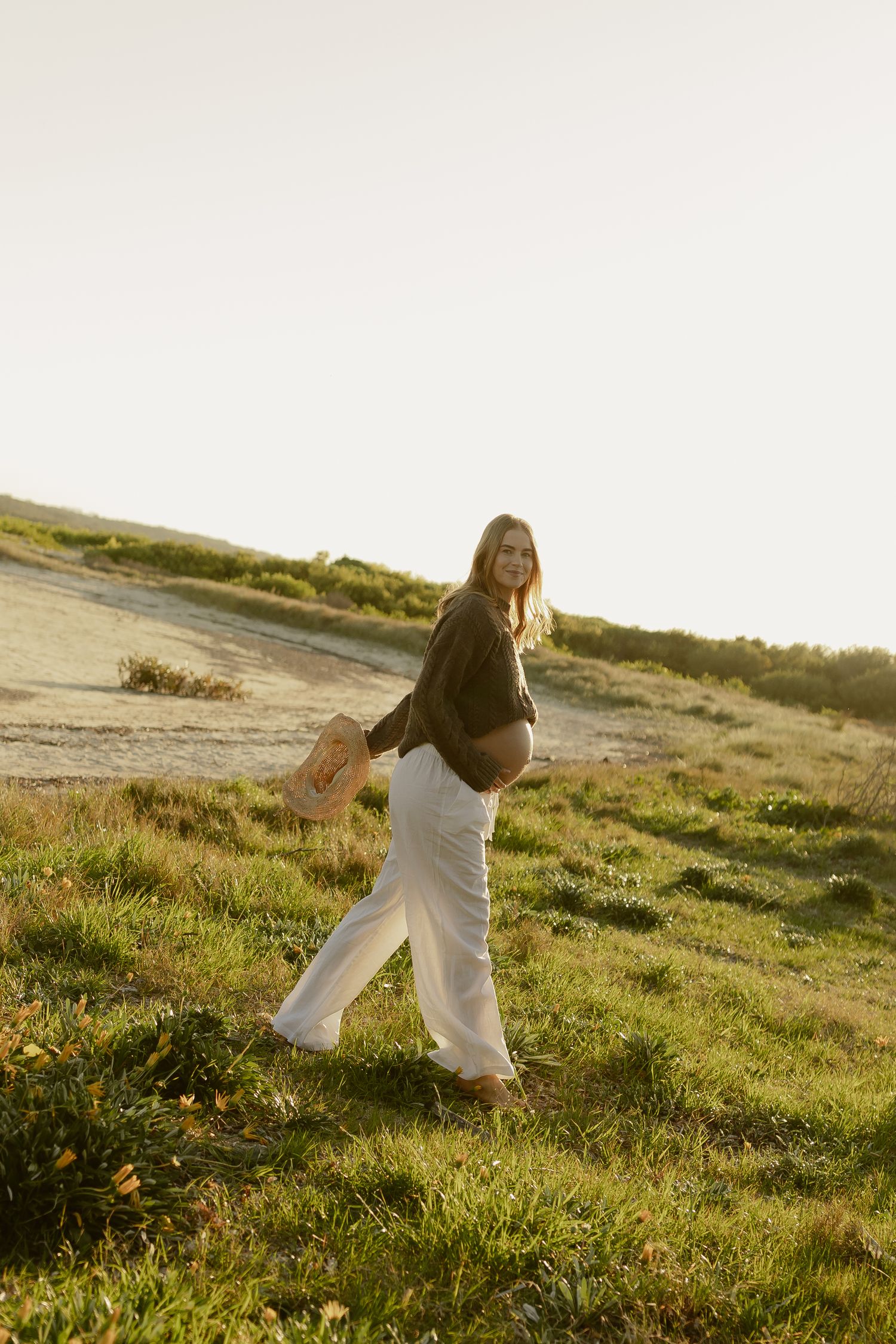 Pregnant woman in white pants and olive top walks on grassy hillside at golden hour holding a straw hat.