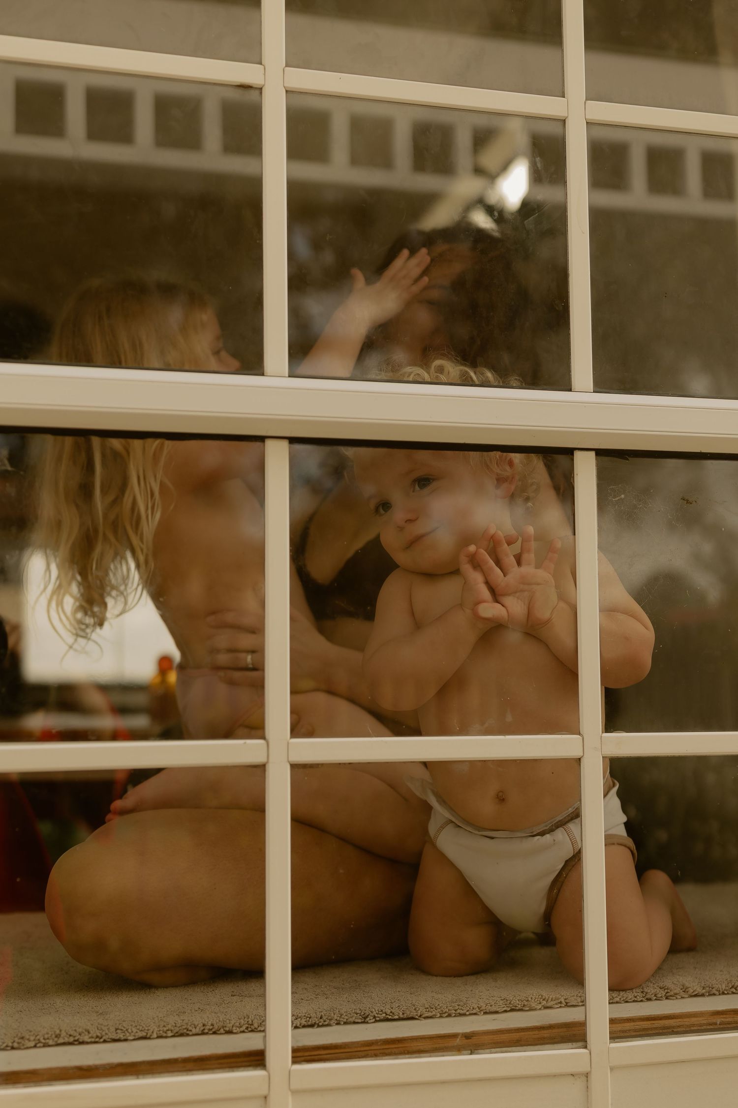 Baby in a diaper sitting on a windowsill pressing hands against the glass with a sibling nearby.