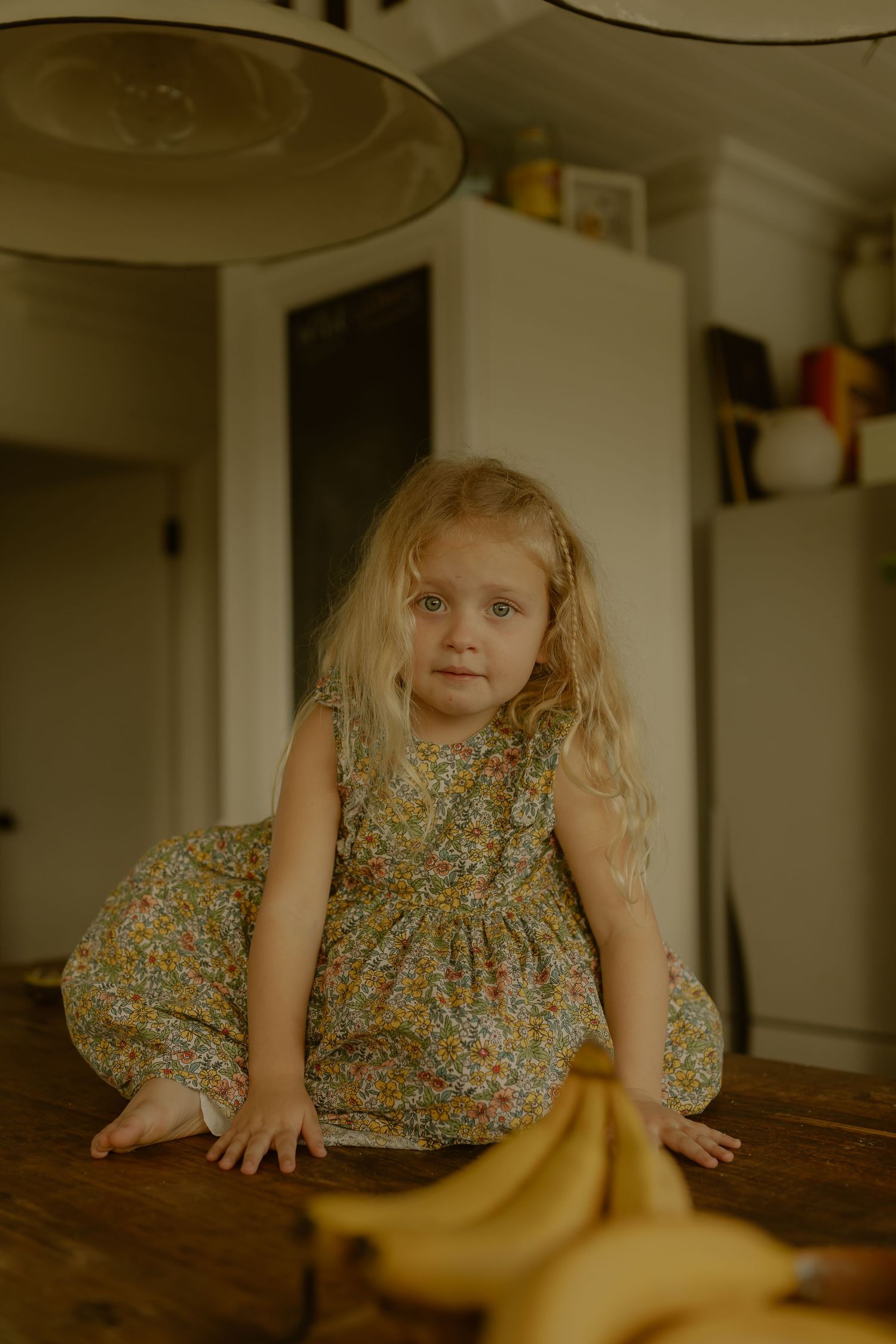 Young blonde girl in floral dress sitting on wooden table near bananas in cozy home kitchen.