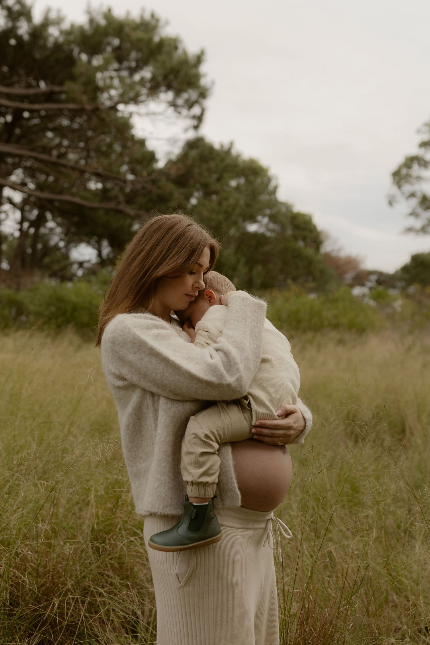 Pregnant woman in beige sweater tenderly embraces toddler son while standing in tall grass outdoors.