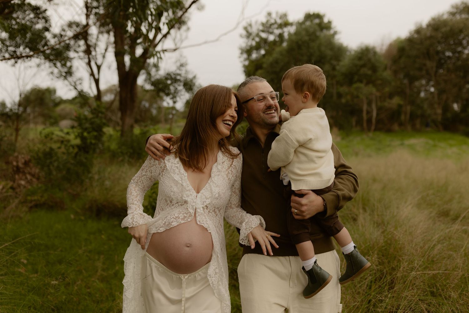 Smiling pregnant woman in white lace dress with partner holding toddler son outdoors in green field.