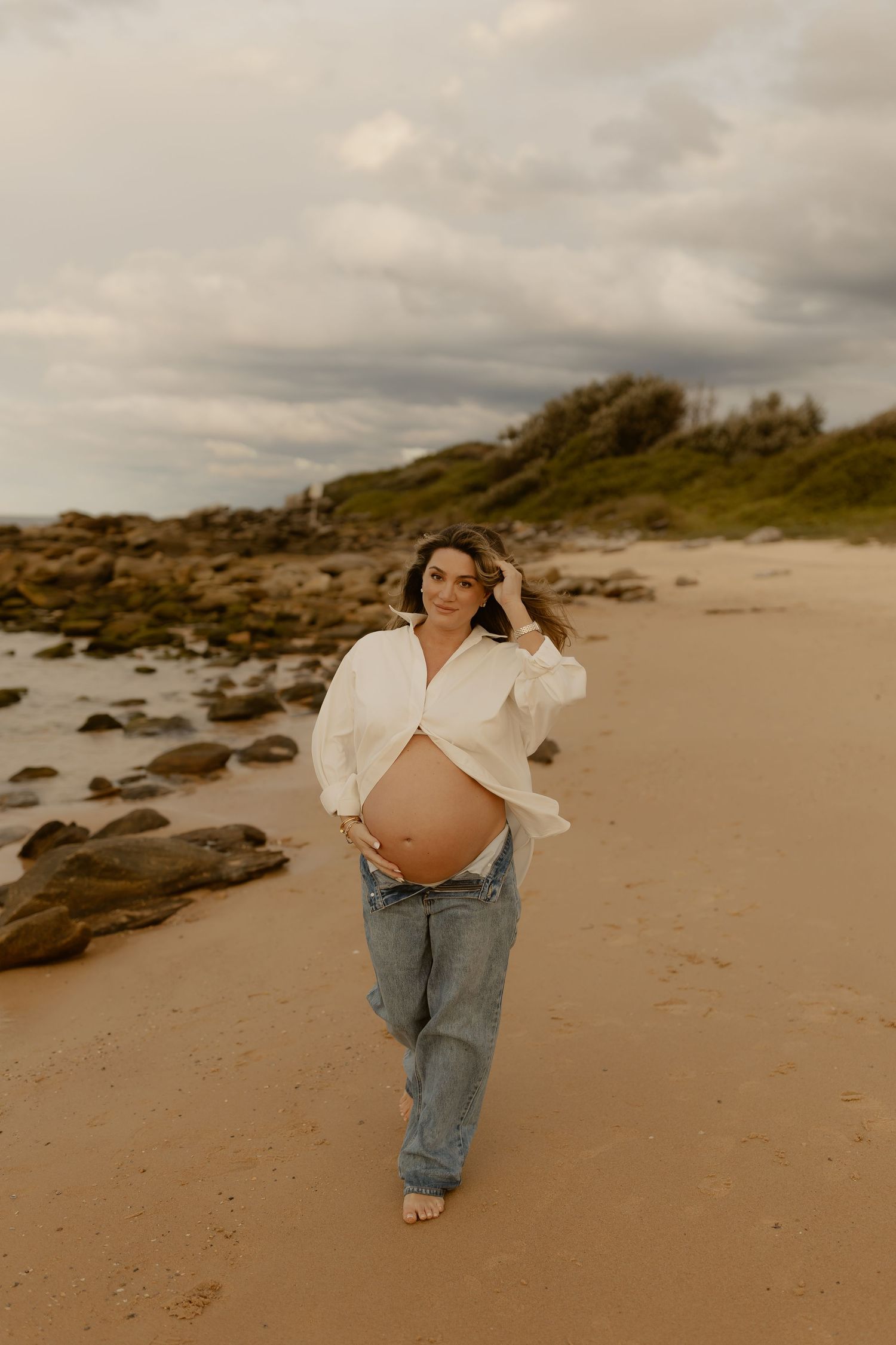 Pregnant woman in white shirt and jeans walking barefoot on sandy beach with rocky coastline.