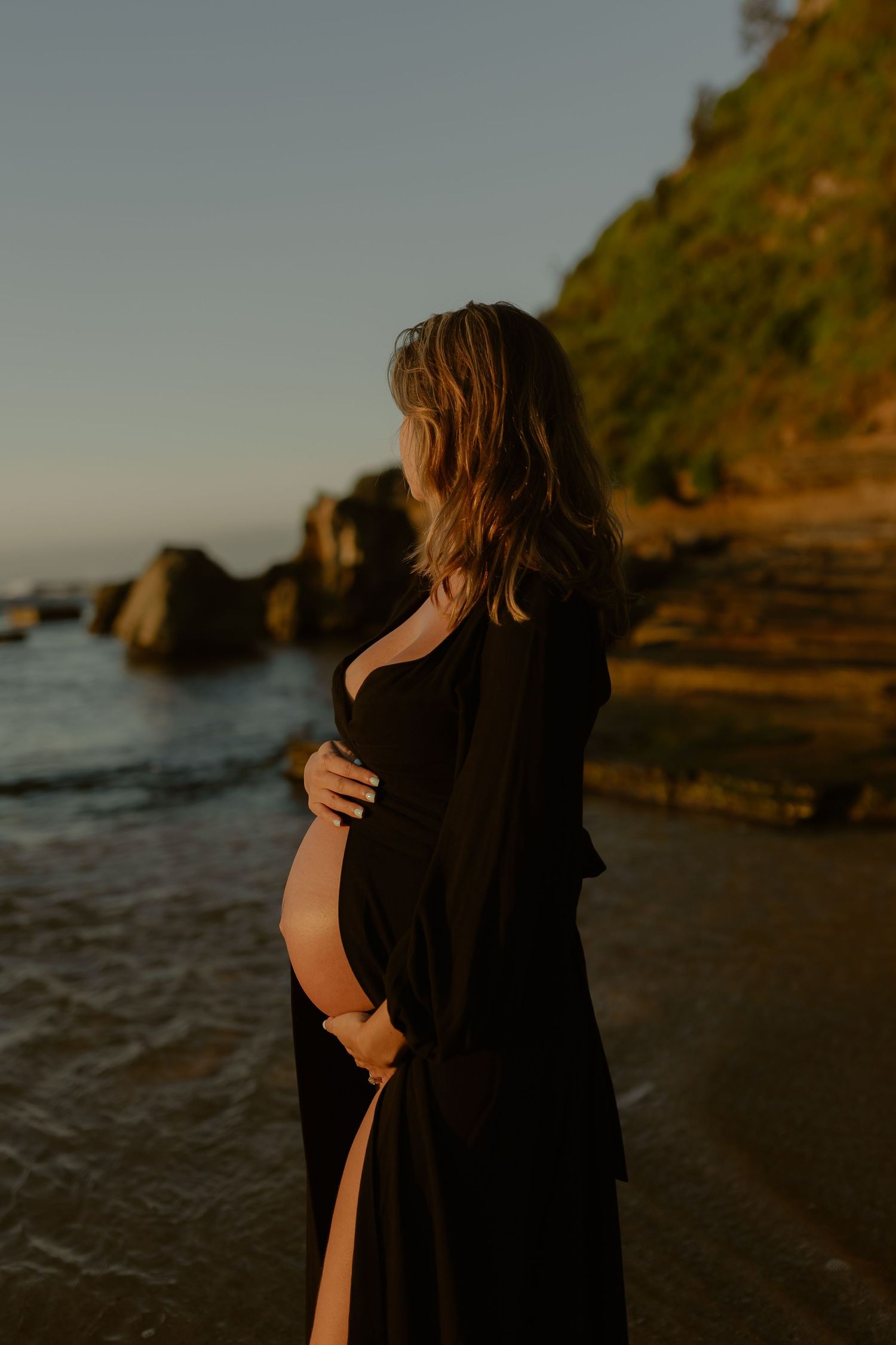 Side profile of pregnant woman in black dress holding belly by ocean rocks at sunset.