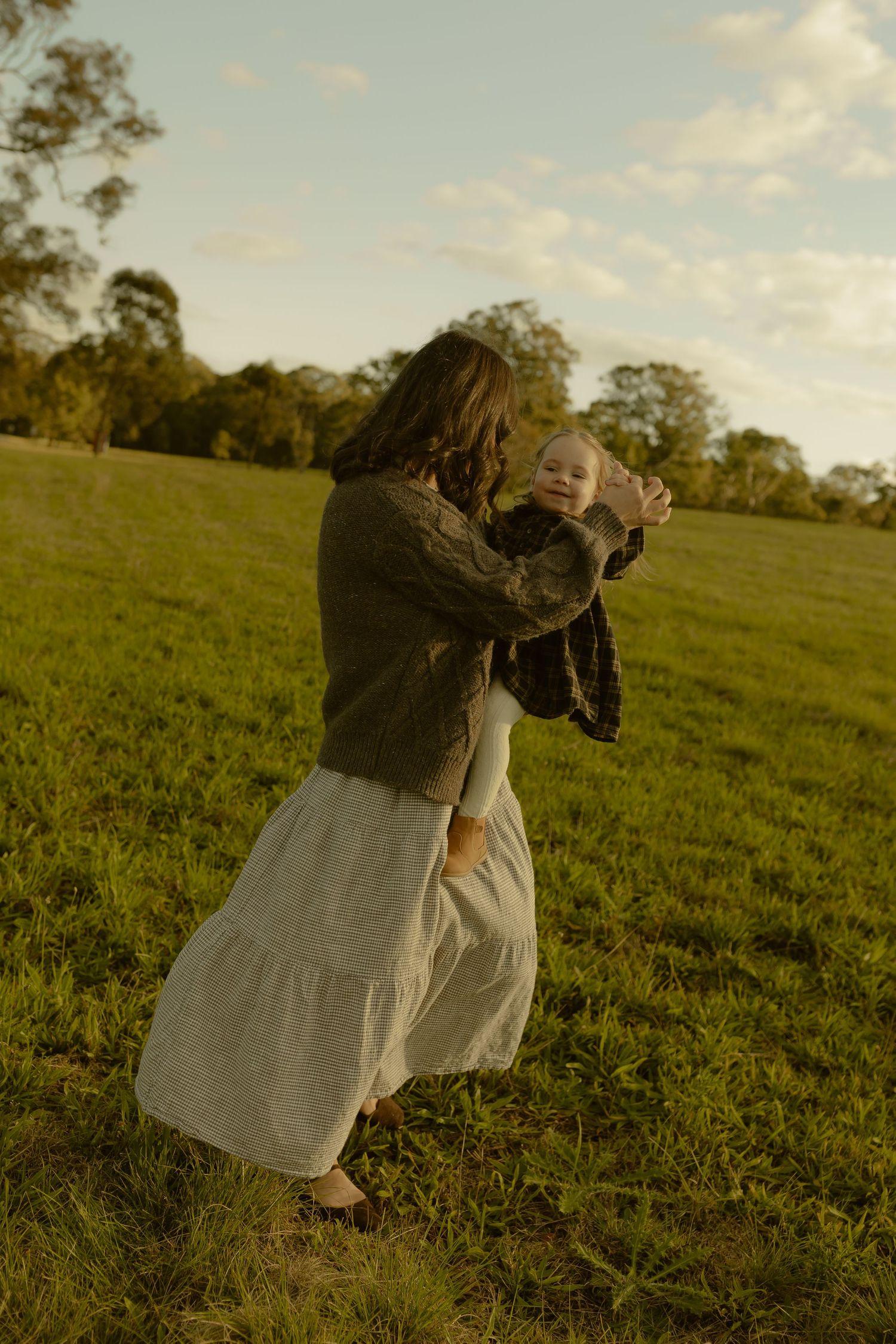 A mother in a white skirt dances with her baby in a sunlit green field with trees in the background.
