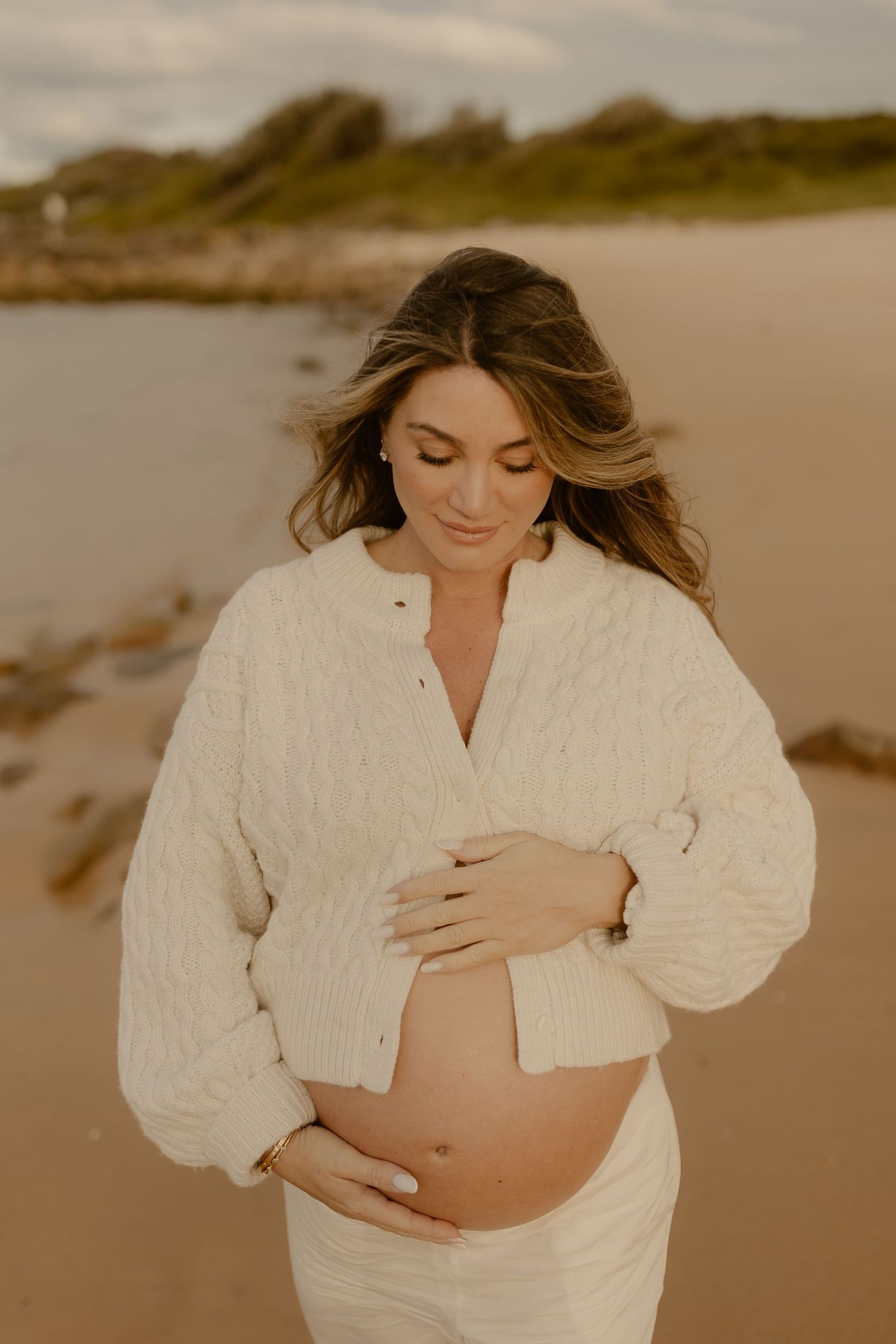 Pregnant woman in cream cable-knit sweater cradling her baby bump on a beach at dusk.