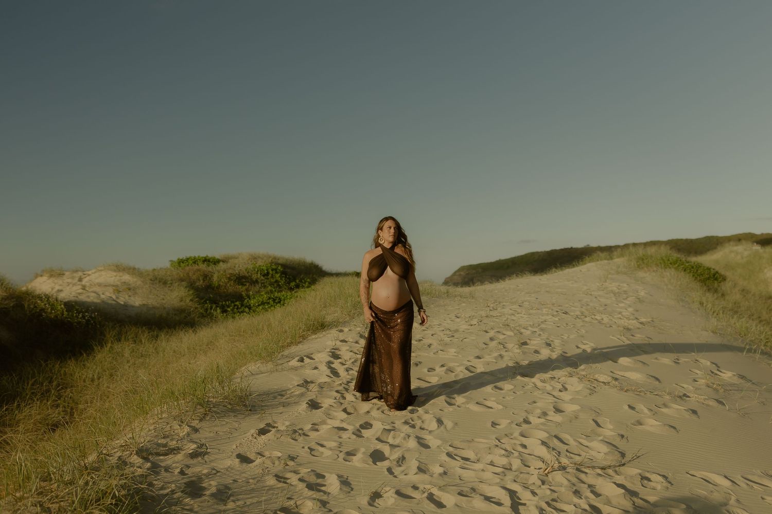 Pregnant woman in brown dress standing on sandy beach path surrounded by dunes and green grass at golden hour.