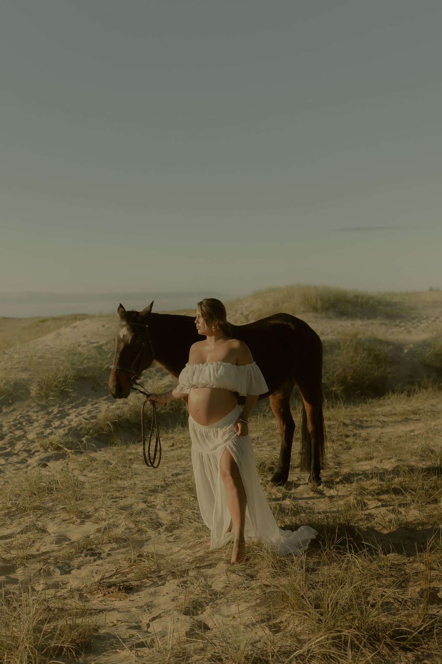 Pregnant woman in white off-shoulder gown stands beside a dark horse on sandy dunes at golden hour.