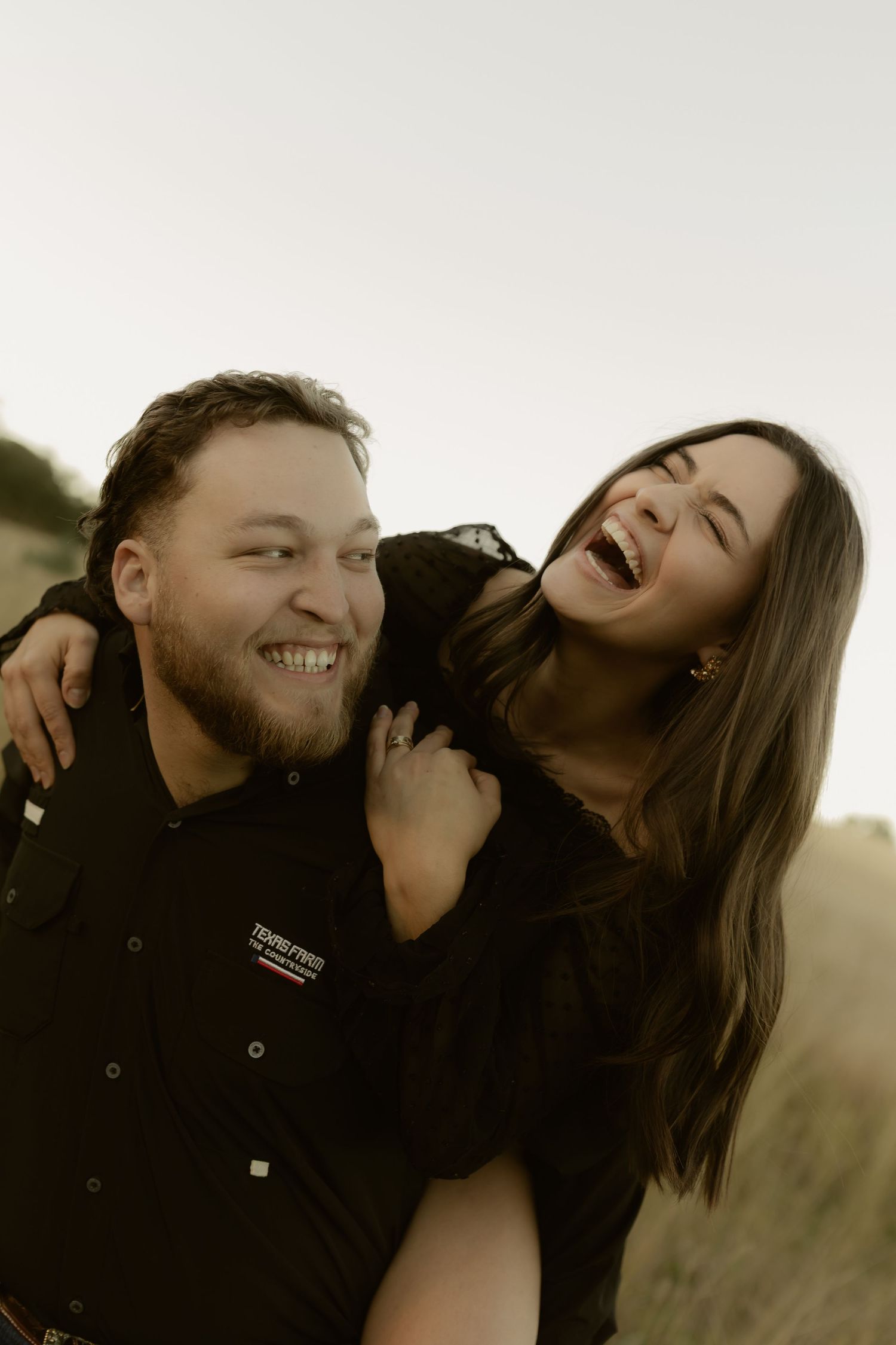A laughing couple sharing a joyful moment outdoors, woman leaning on mans shoulders smiling.